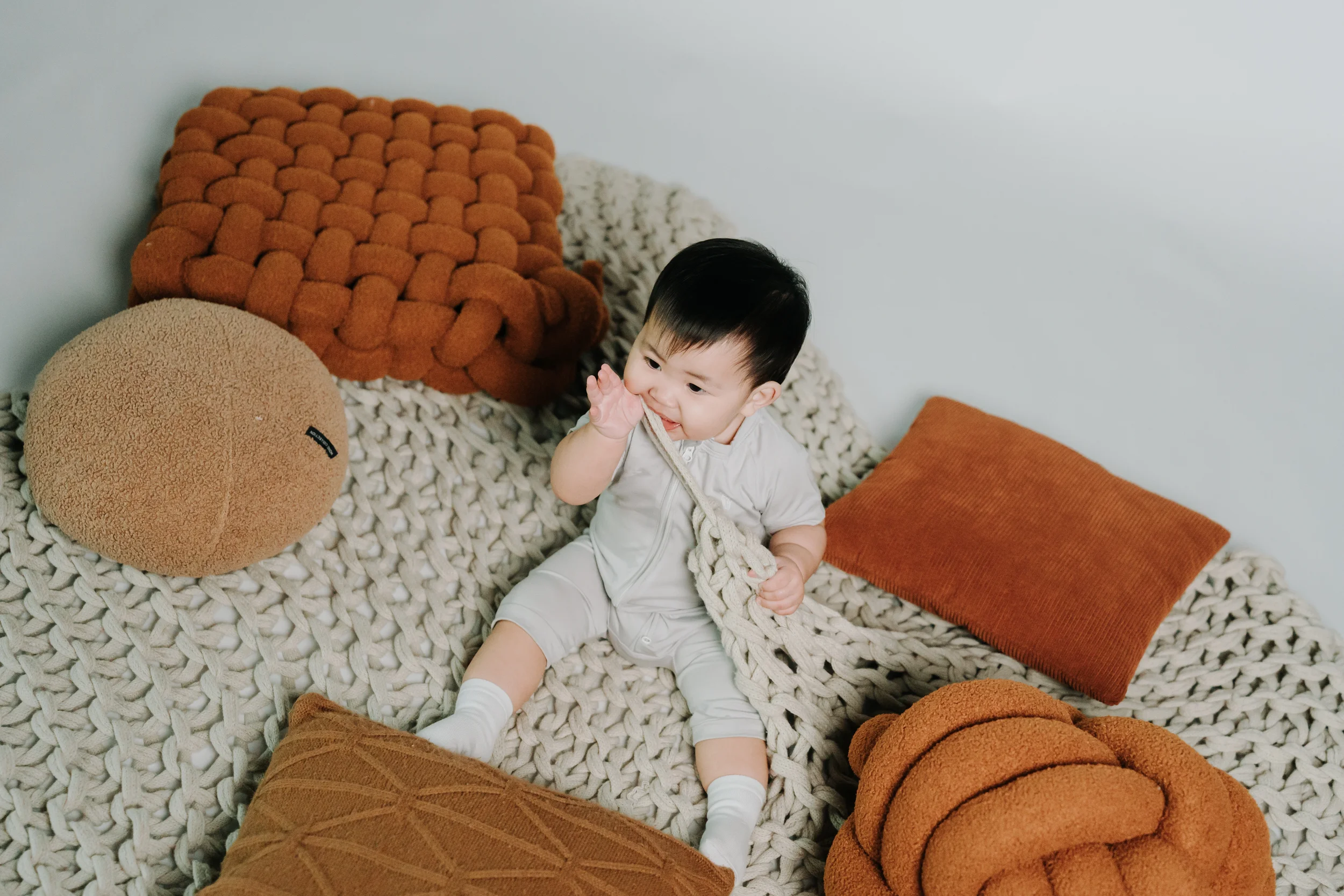 Cute baby sitting on a chunky knit blanket surrounded by decorative pillows in shades of orange and brown.