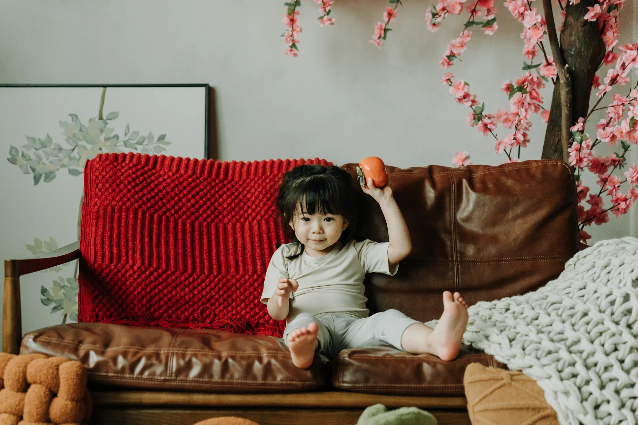 Young girl sitting on a brown leather couch with a red blanket behind her, holding a orange apple in her raised right hand, next to a white chunky knit blanket, with pink blossom branch in the background.