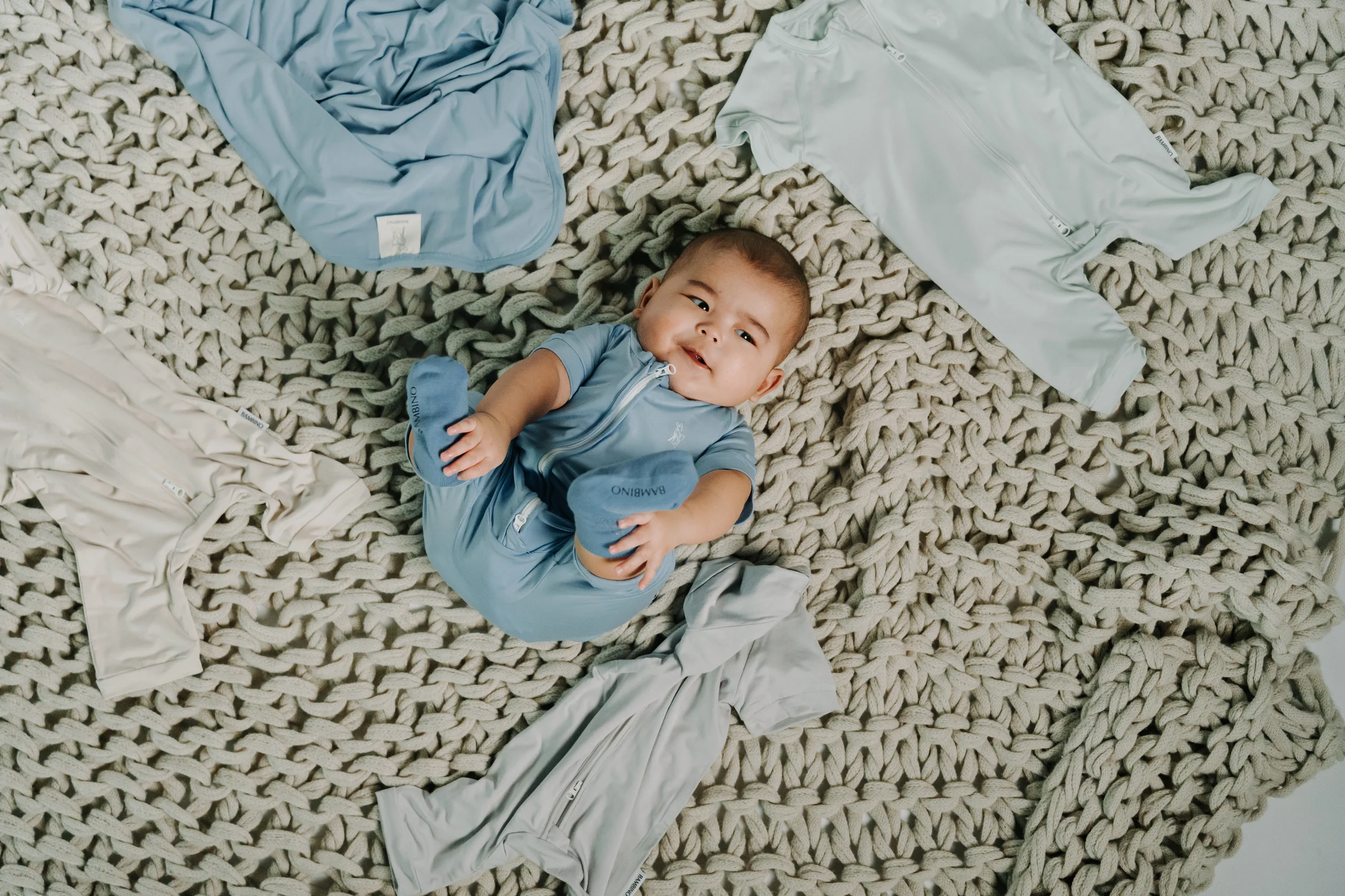 A smiling baby in a blue outfit lying on a chunky knit beige blanket surrounded by various light-colored clothing items.
