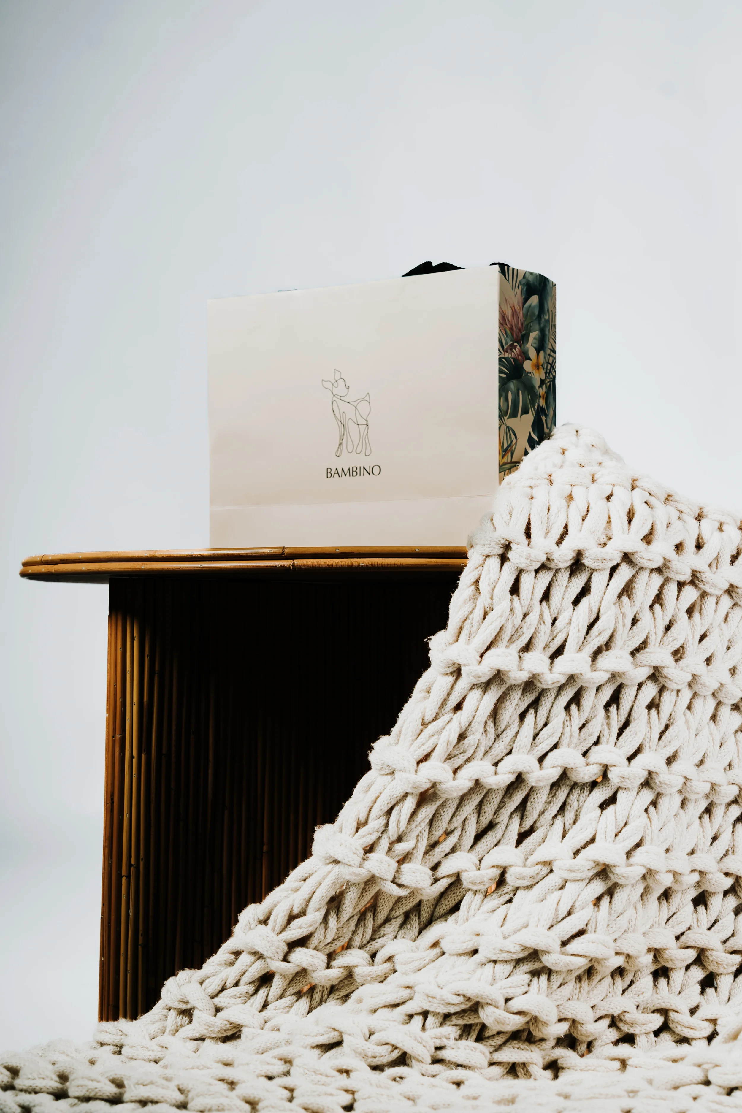 A paper shopping bag with a minimalist deer logo and the text 'BAMBINO' on a wooden table, with a chunky off-white knitted blanket draped nearby.