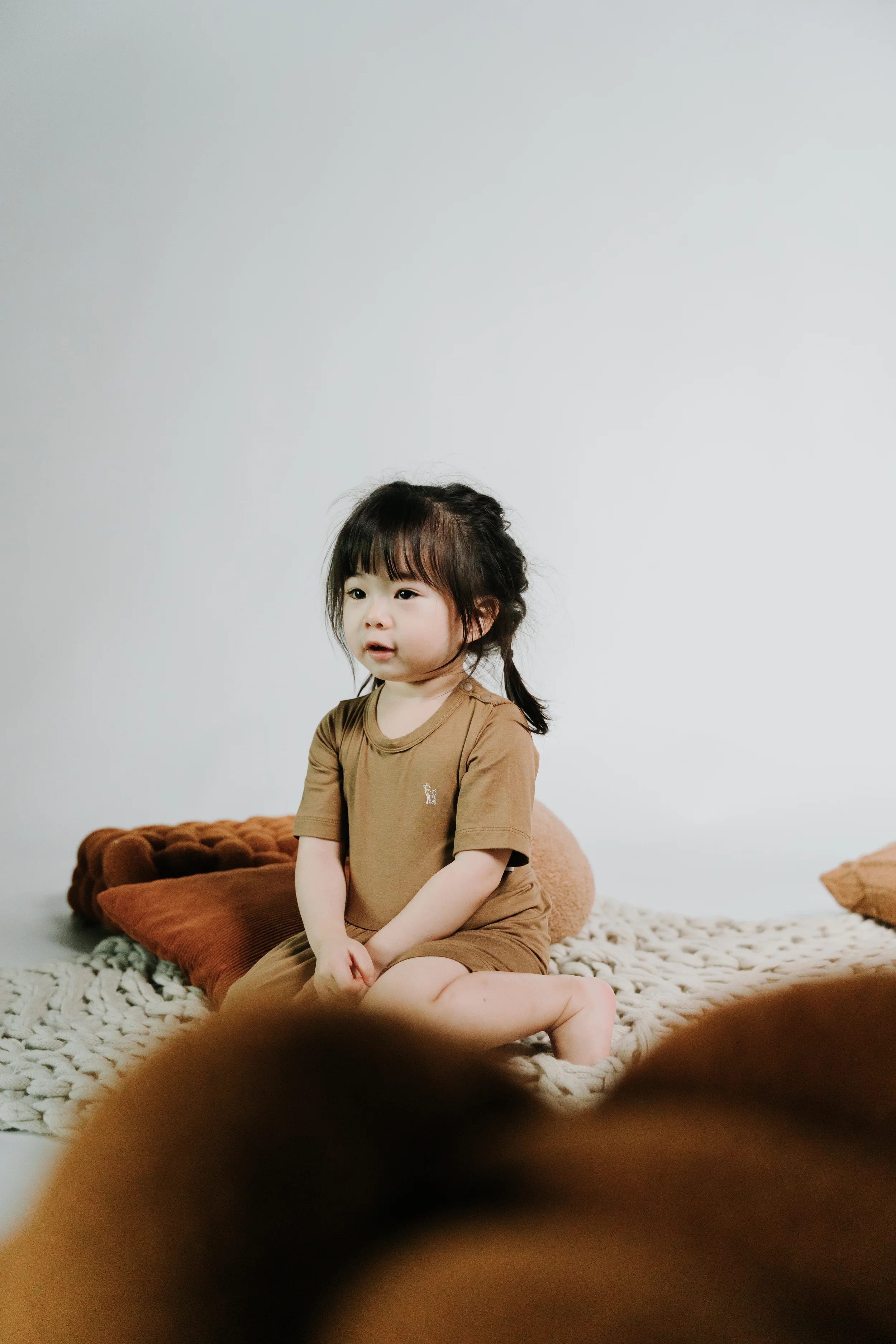 A young girl with dark hair sitting on a textured white blanket, wearing a brown shirt and shorts, with a neutral expression, against a plain white wall.