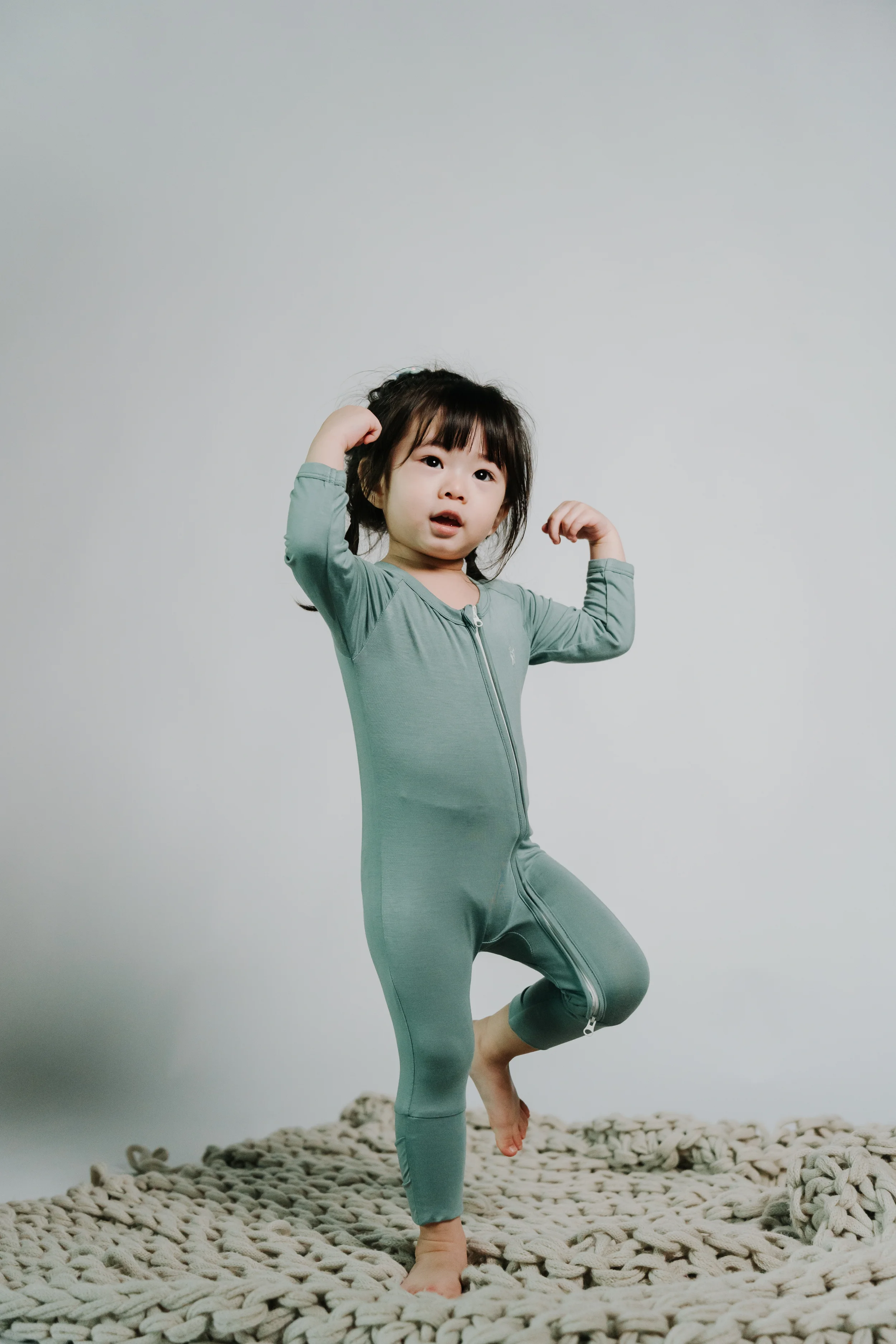 A young girl with dark hair and bangs wearing a green jumpsuit, standing on a chunky knit rug against a plain gray background, striking a playful pose with her arms flexed.