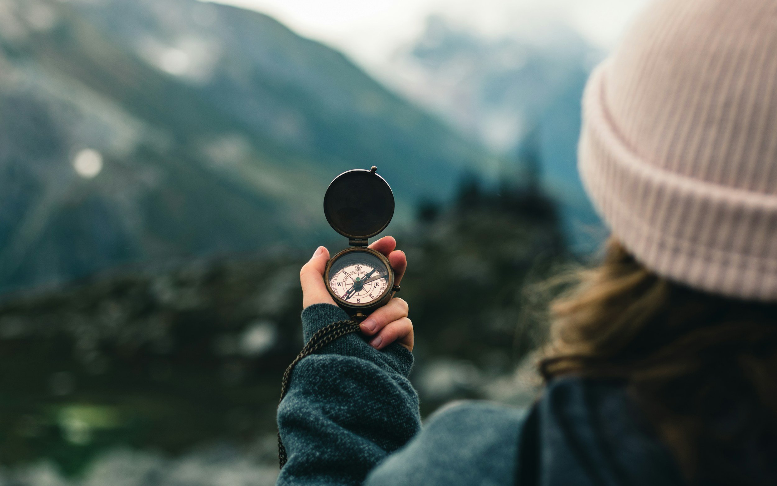 A person holding a compass outdoors with mountains and sky in the background.