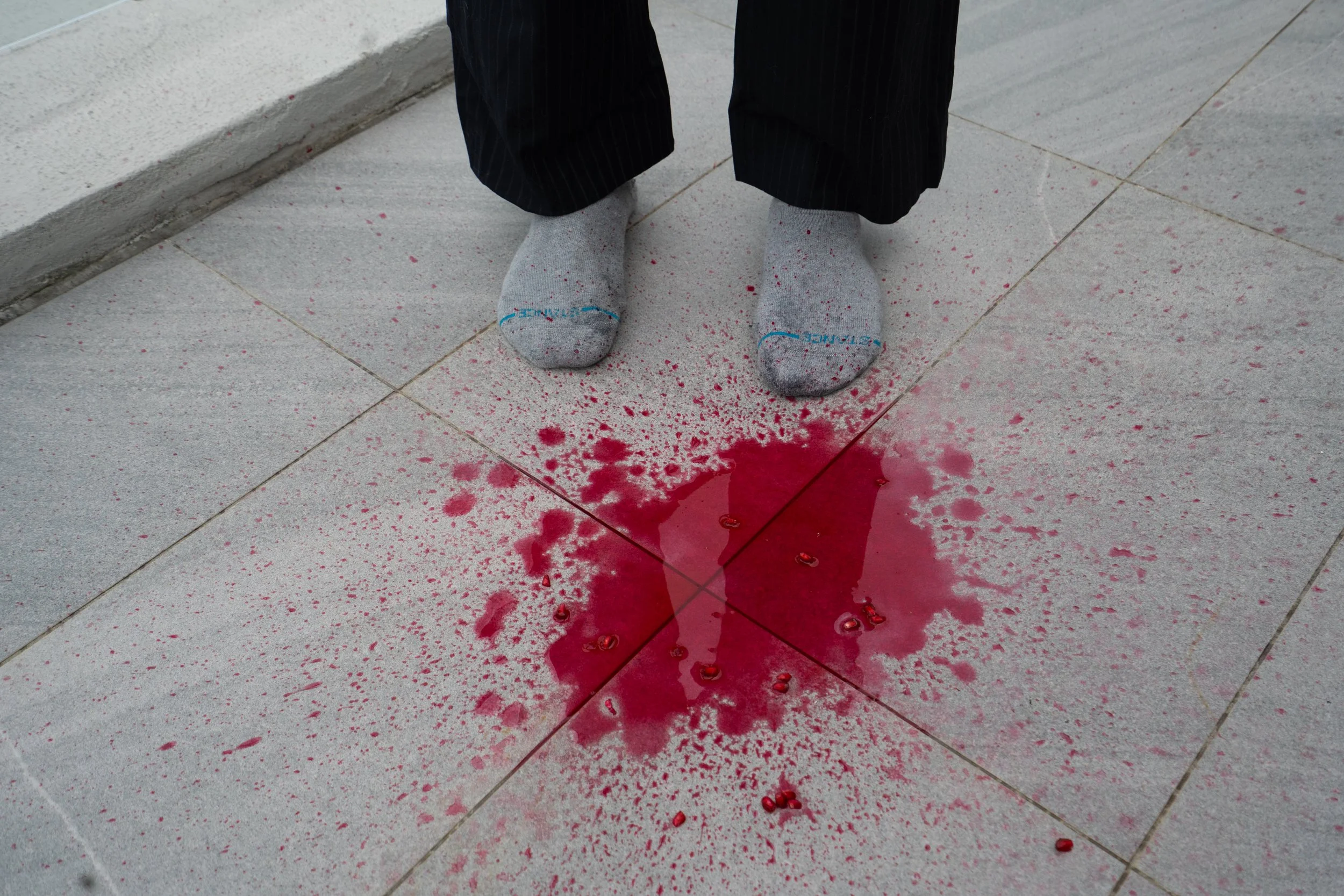 Person standing on tiled floor with red spilled liquid and blood-like droplets, wearing gray socks and black pants.