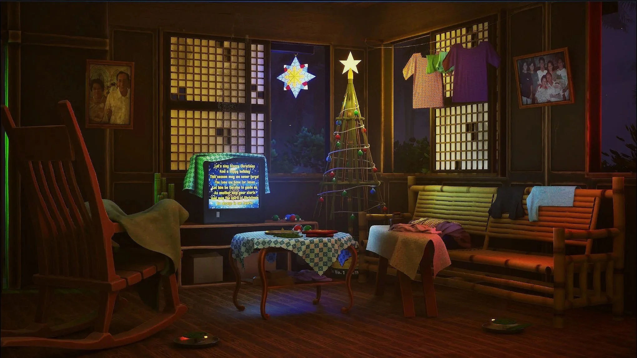 A cozy living room decorated for Christmas with a small, lit Christmas tree, a TV displaying text, and holiday clothes hanging to dry. There are wooden chairs, a table with a patterned tablecloth, framed family photos on the wall, and a window showin