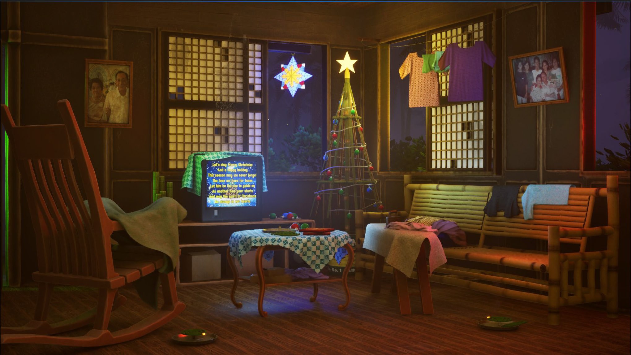 Living room decorated for Christmas with a small illuminated Christmas tree, a television with Christmas music, a rocking chair, a bamboo sofa with blankets, a small table with a patterned cloth, clothes hanging to dry, and family photos on the walls.