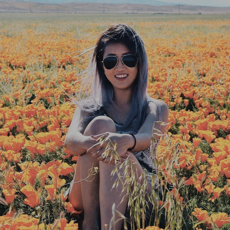 A young woman with sunglasses and long hair sitting in a field of orange flowers, smiling at the camera.