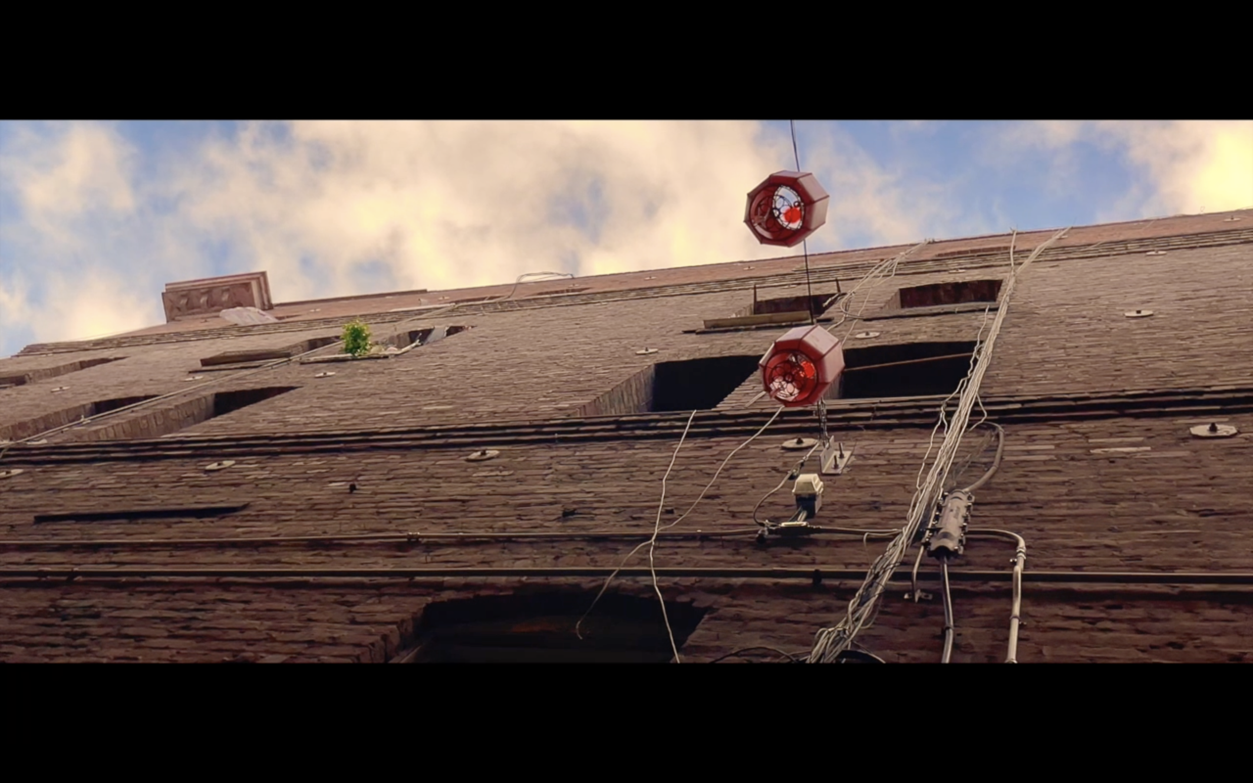 Low-angle view of a brick building with two red warning lights and various wires on the exterior, against a partly cloudy sky.