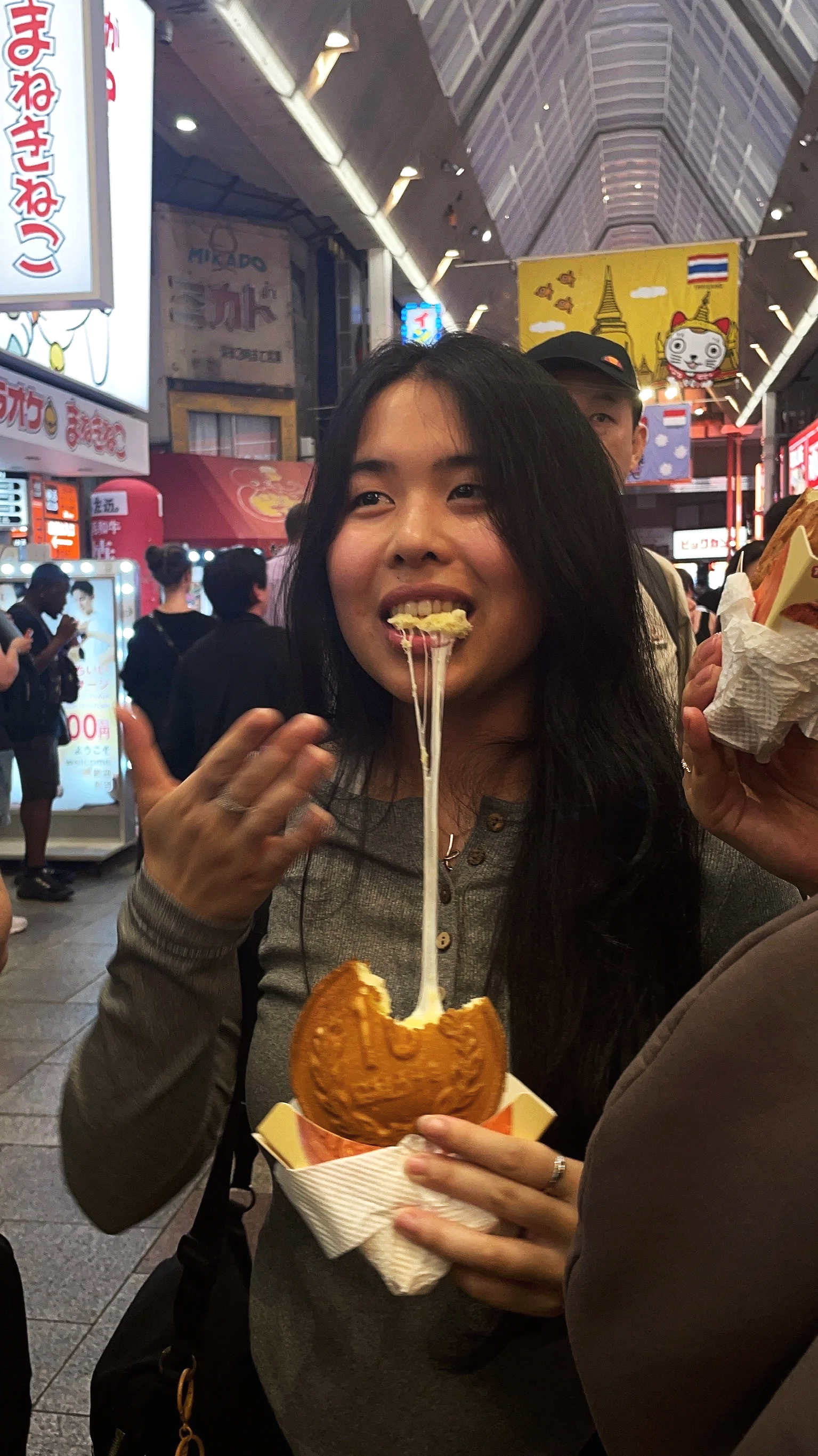 A woman with long black hair eating cheese-filled taiyaki, a fish-shaped pastry, in a busy indoor market.
