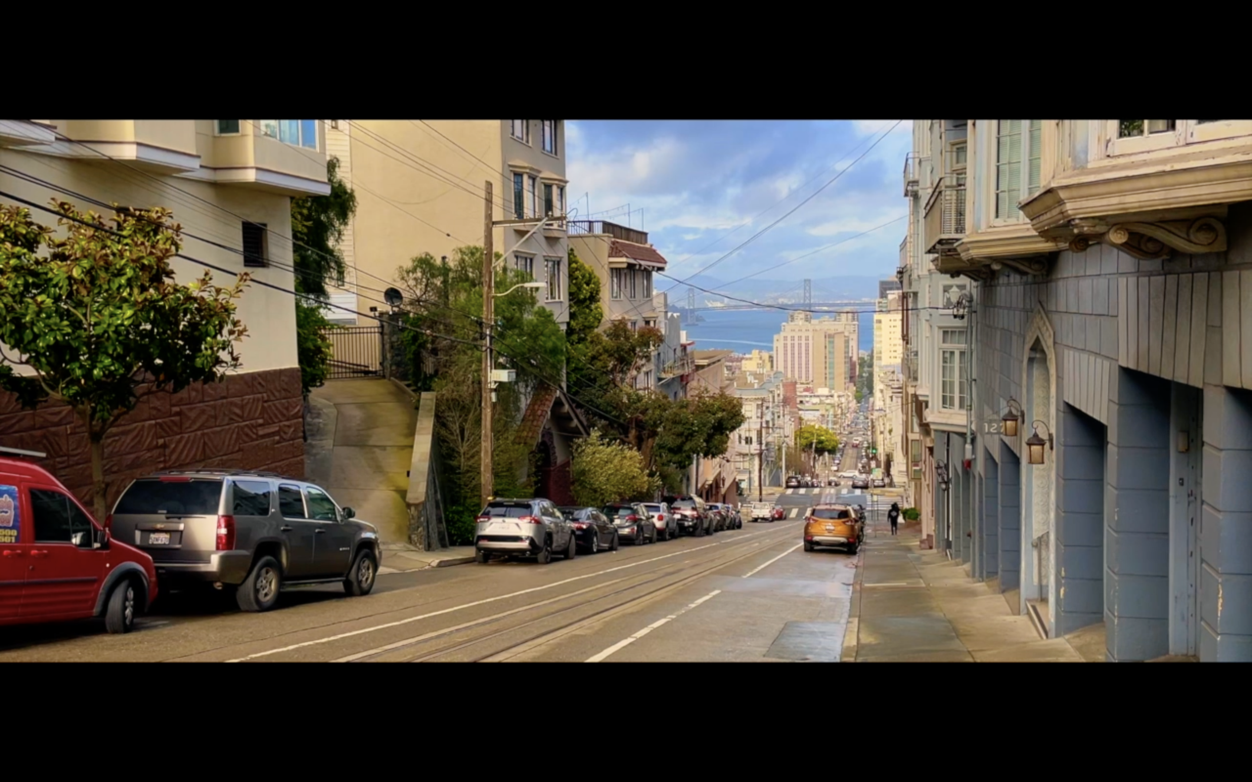 A city street view with parked cars, residential buildings, trees, and a distant view of a bridge over water.