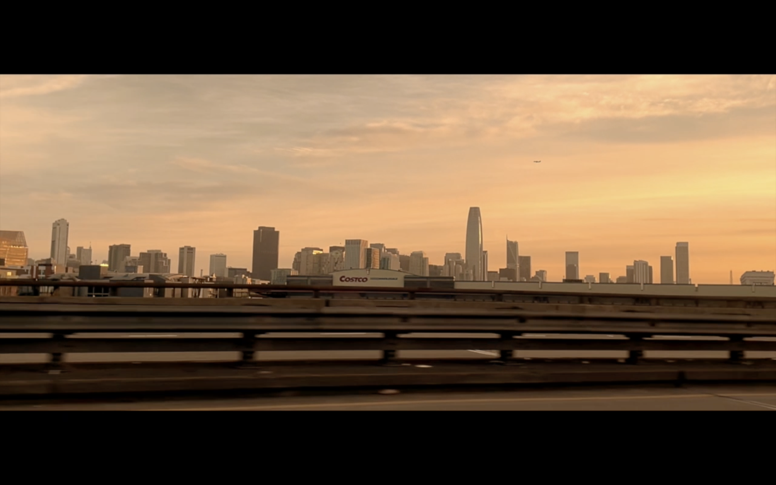 Sunset view of San Francisco skyline with skyscrapers, including Salesforce Tower and Salesforce Park, seen from a highway with guardrails in the foreground, and a Costco sign visible. The sky is painted with soft orange and pink hues, and an airplan