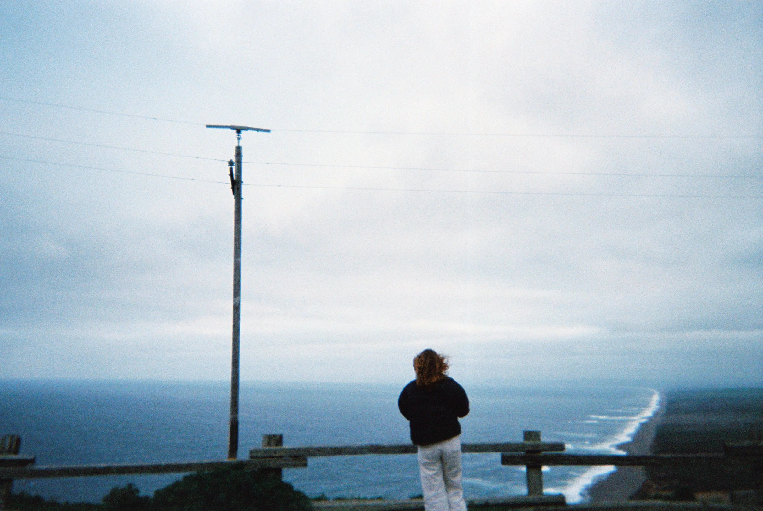 A person with curly hair standing on a lookout, facing a large body of water with cliffs, under a cloudy sky.