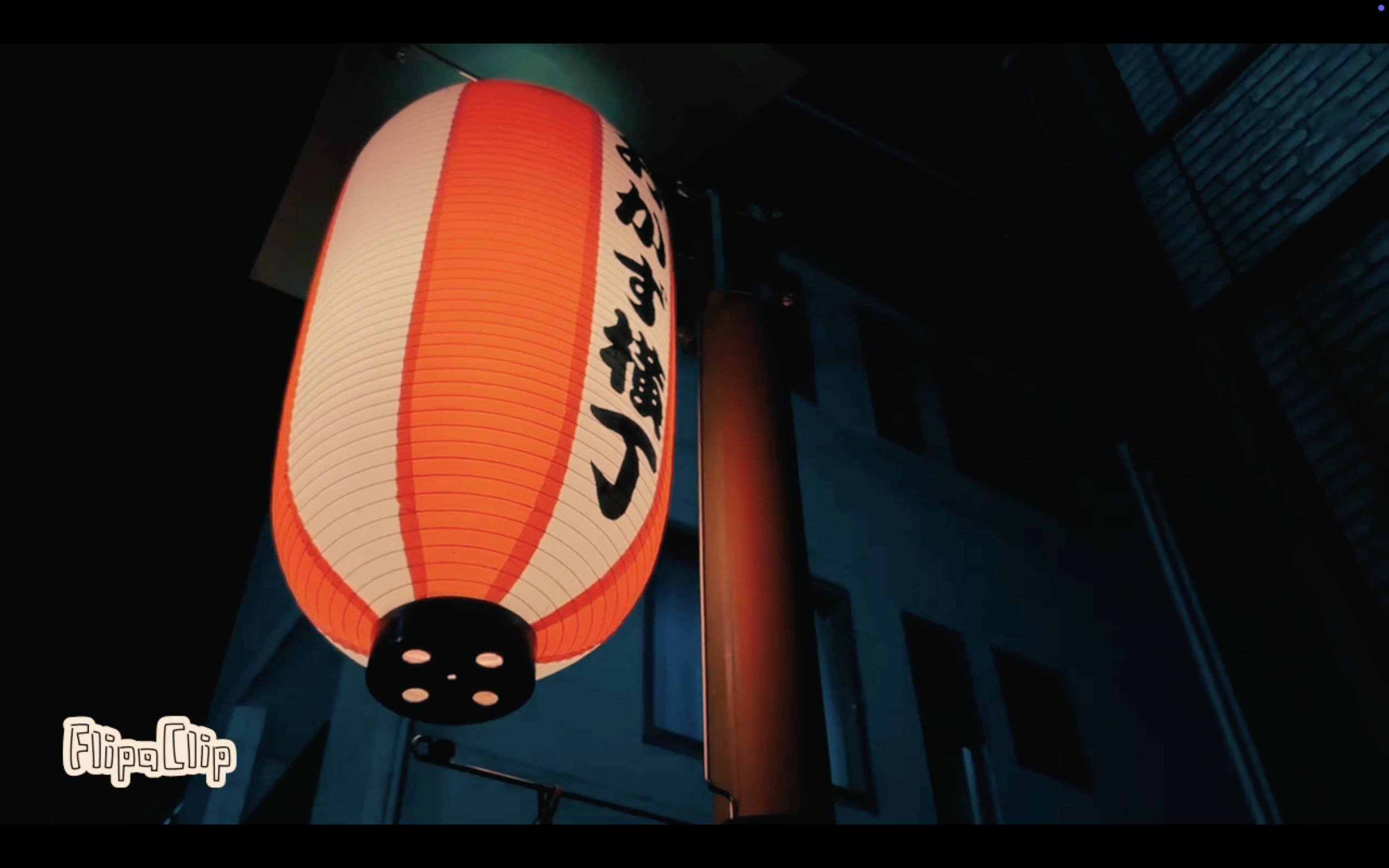 A traditional Japanese paper lantern lit up at night, hanging outdoors, with black Japanese characters on its surface.