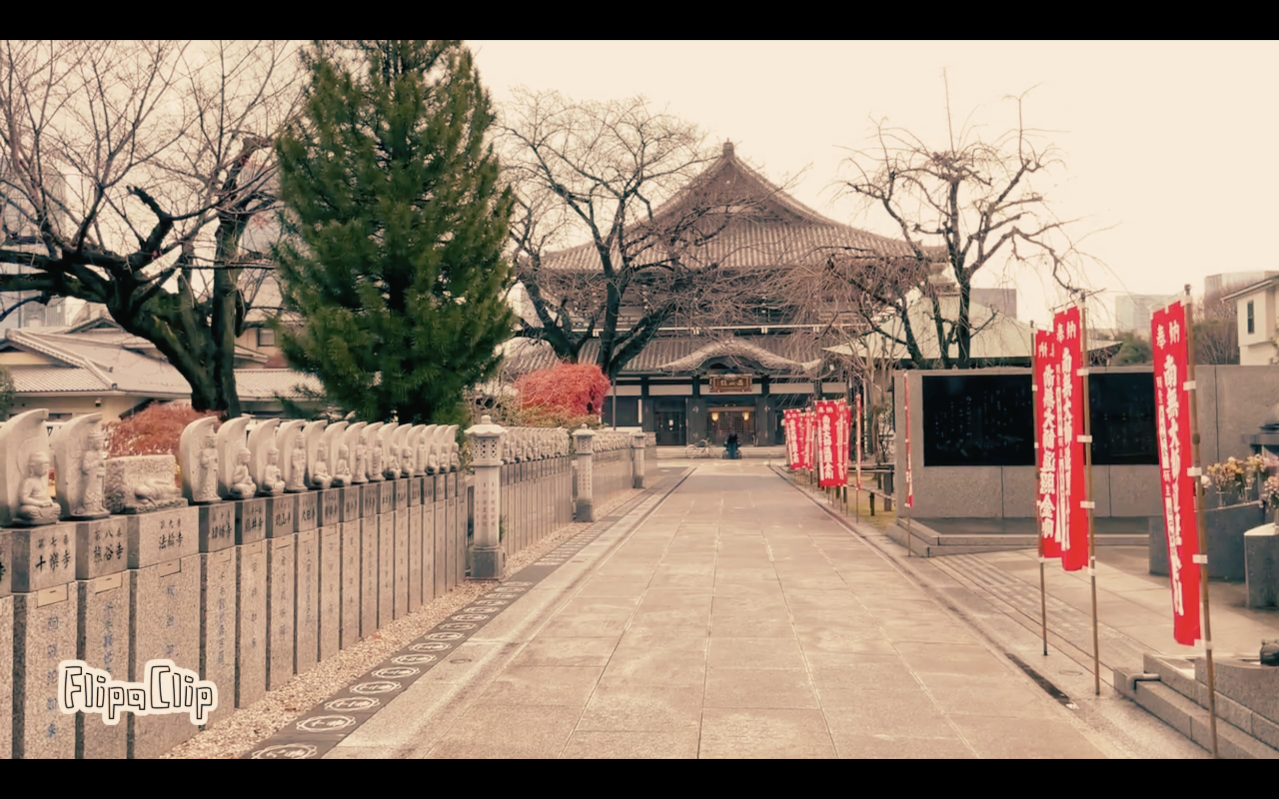 Path leading to a traditional Japanese temple, lined with stone statues and red banners, with leafless trees and some green foliage.