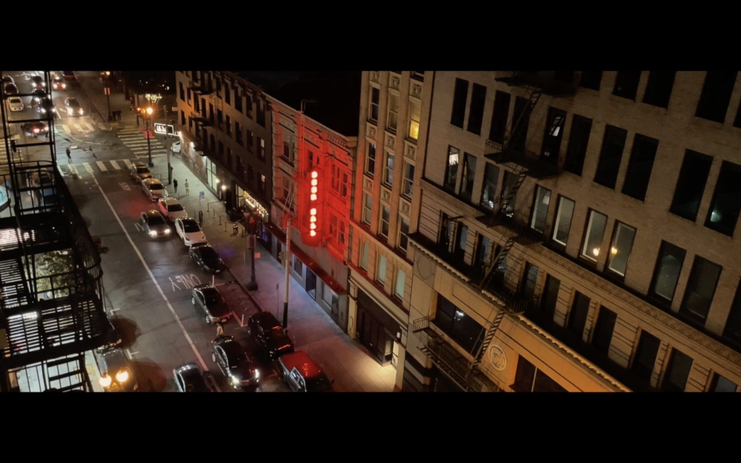 Nighttime city street view with parked cars, illuminated storefronts, and a building with a red neon sign.