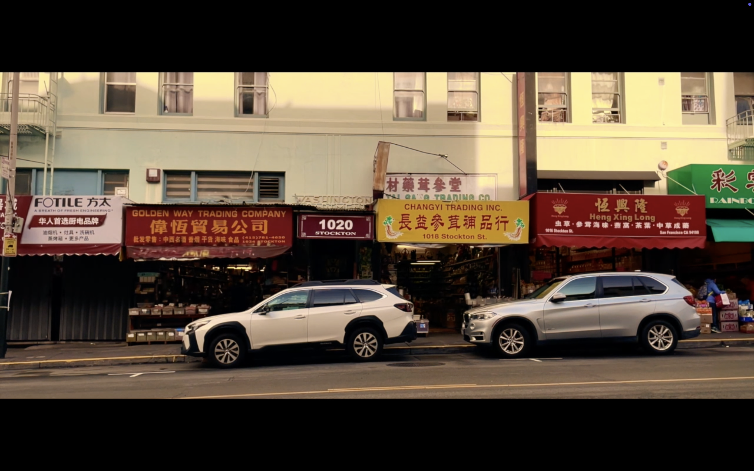 Street view with storefronts featuring Chinese signs, parked white and silver SUVs on the street.