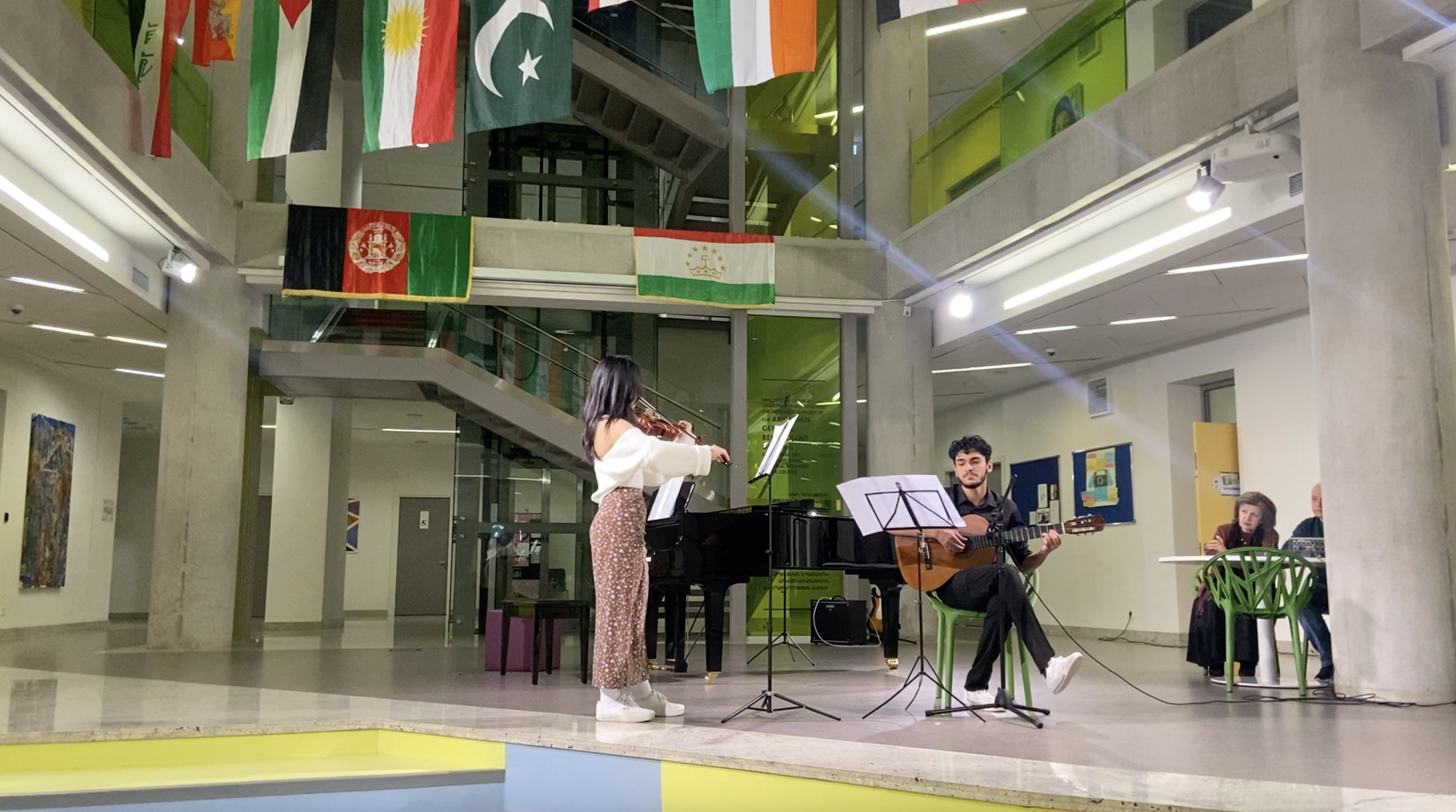 A musical performance in a modern space with flags hanging from the ceiling. A girl playing a violin and a young man playing a guitar are on stage, while an audience watches from tables.