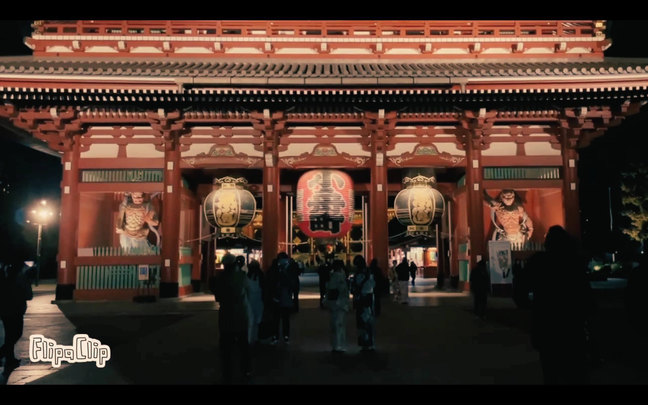 Night view of a traditional Japanese temple gate with large hanging lanterns and samurai warrior posters, people walking underneath.