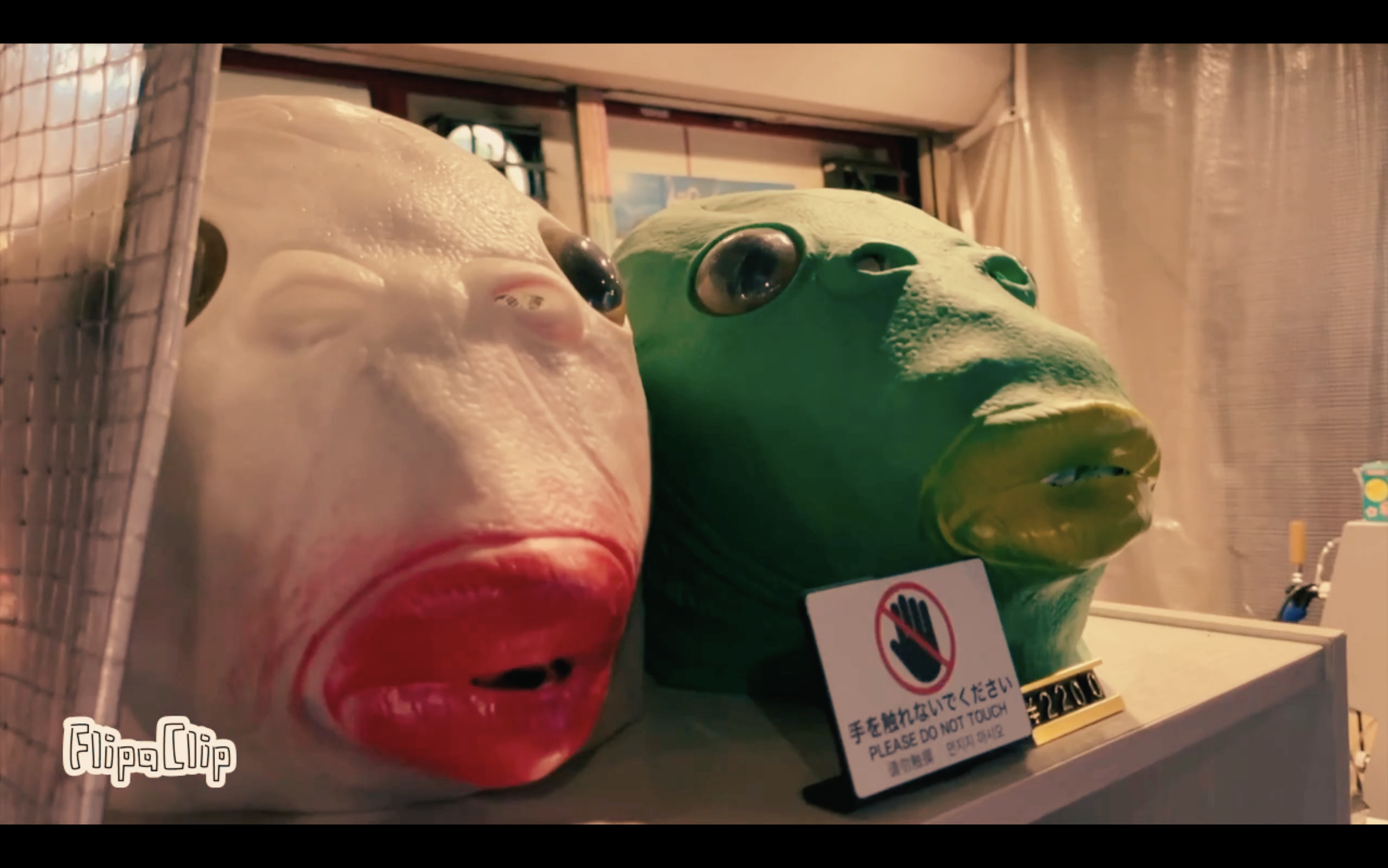 Two large, colorful masks—one resembling a face with exaggerated red lips and another resembling a frog—on display, with a sign that says "Please do not touch" in English and Japanese.