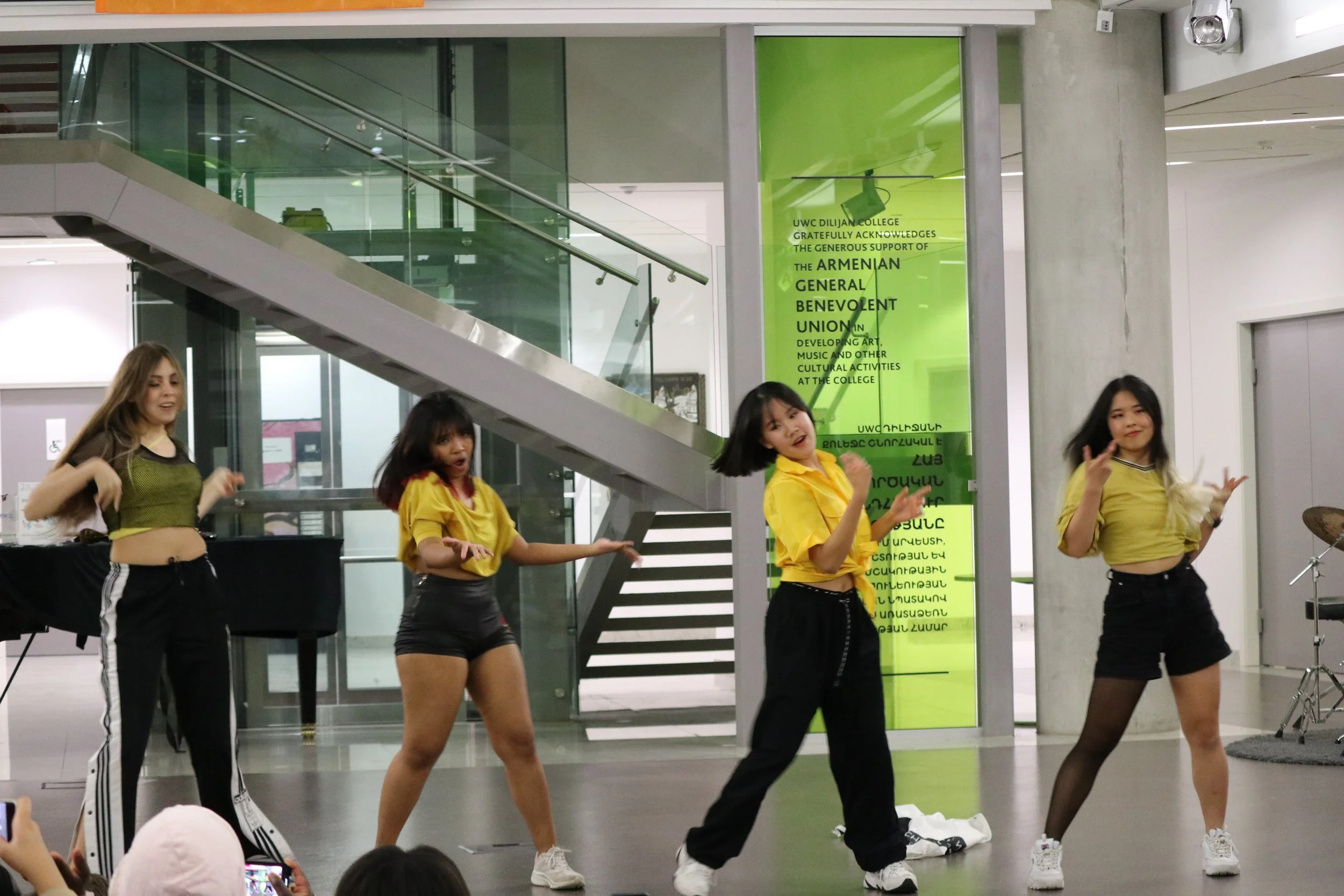 Four young women are performing a dance on a stage inside a modern building. They are all wearing yellow and black outfits. An audience is visible in the foreground, taking pictures.