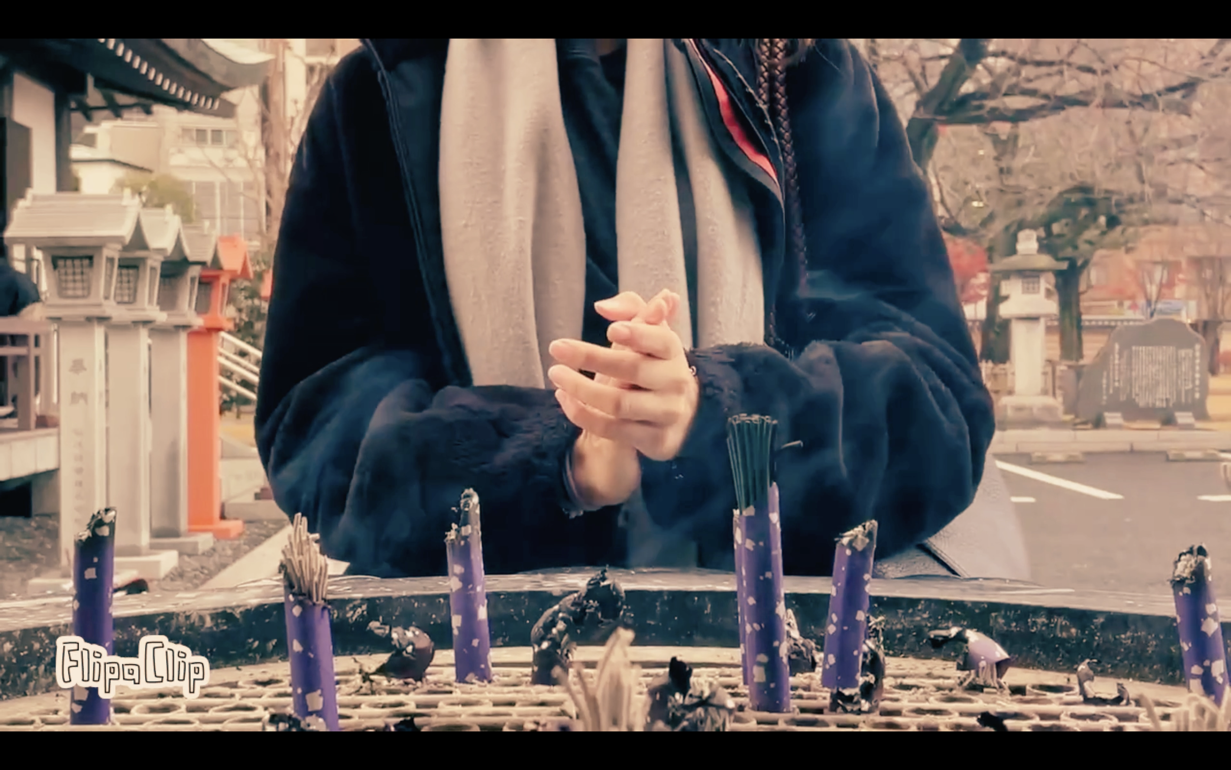 A person standing outdoors near a shrine or memorial, with hands clasped in prayer or contemplation, behind a container with burnt incense sticks and ashes.