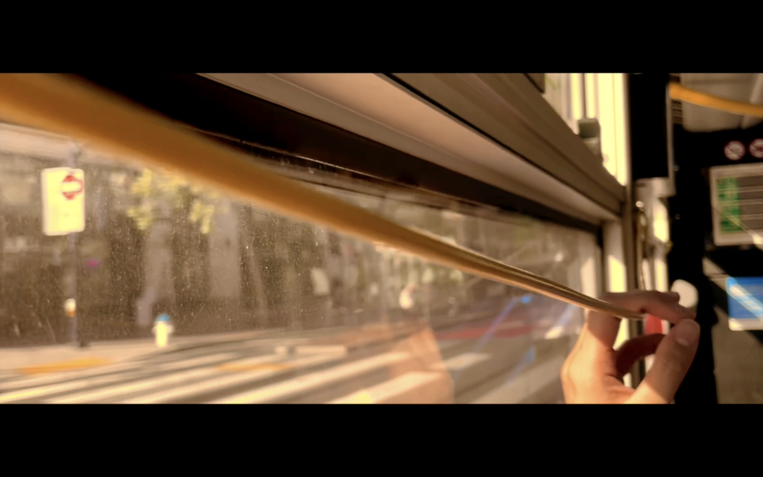 A person holds onto a horizontal metal handrail on a bus or train, with a view of the outside city street through a window, reflecting sunlight.
