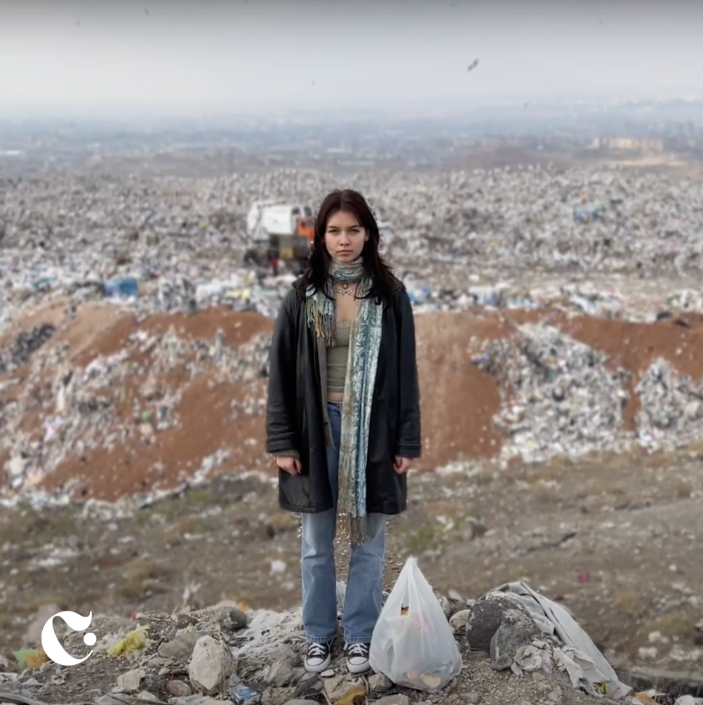 A young woman stands on a street amidst a large landfill of trash and debris in a cityscape background.