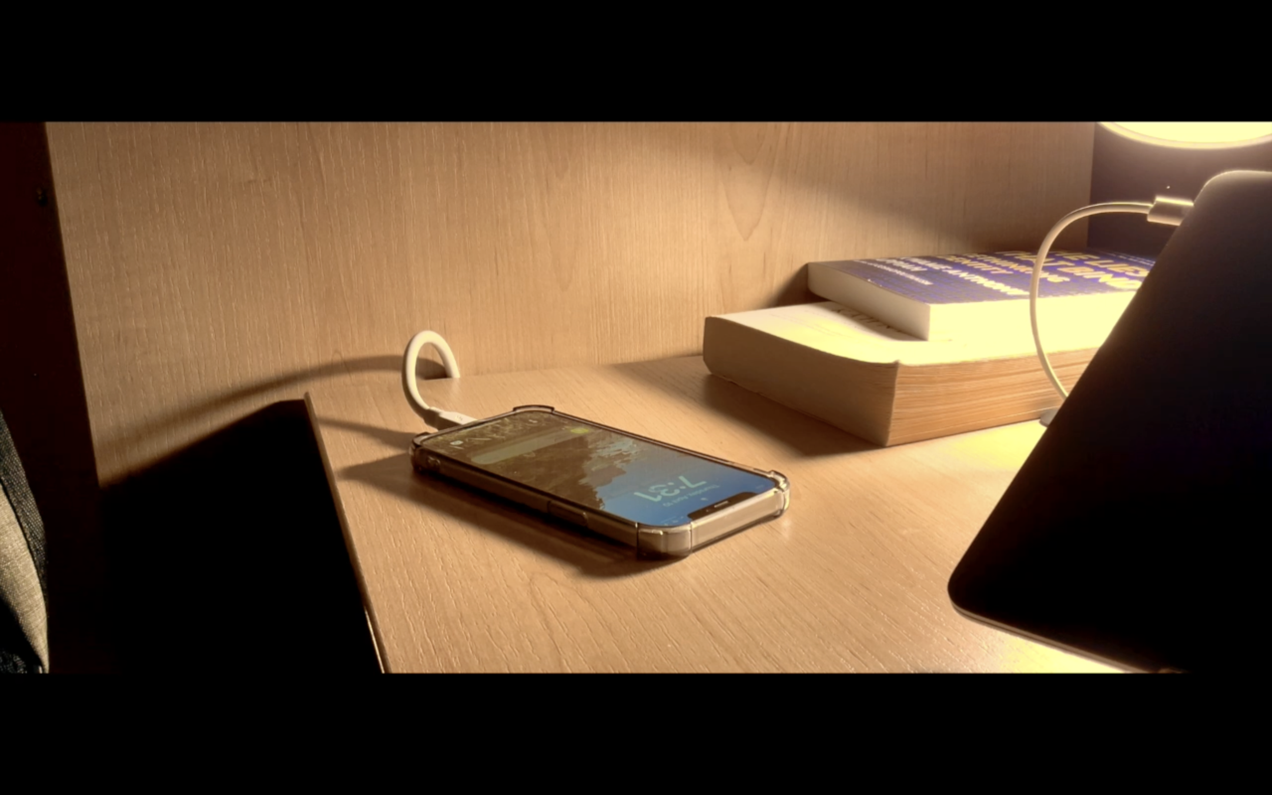 A wooden bedside table with a smartphone, a stack of books, and a lamp.