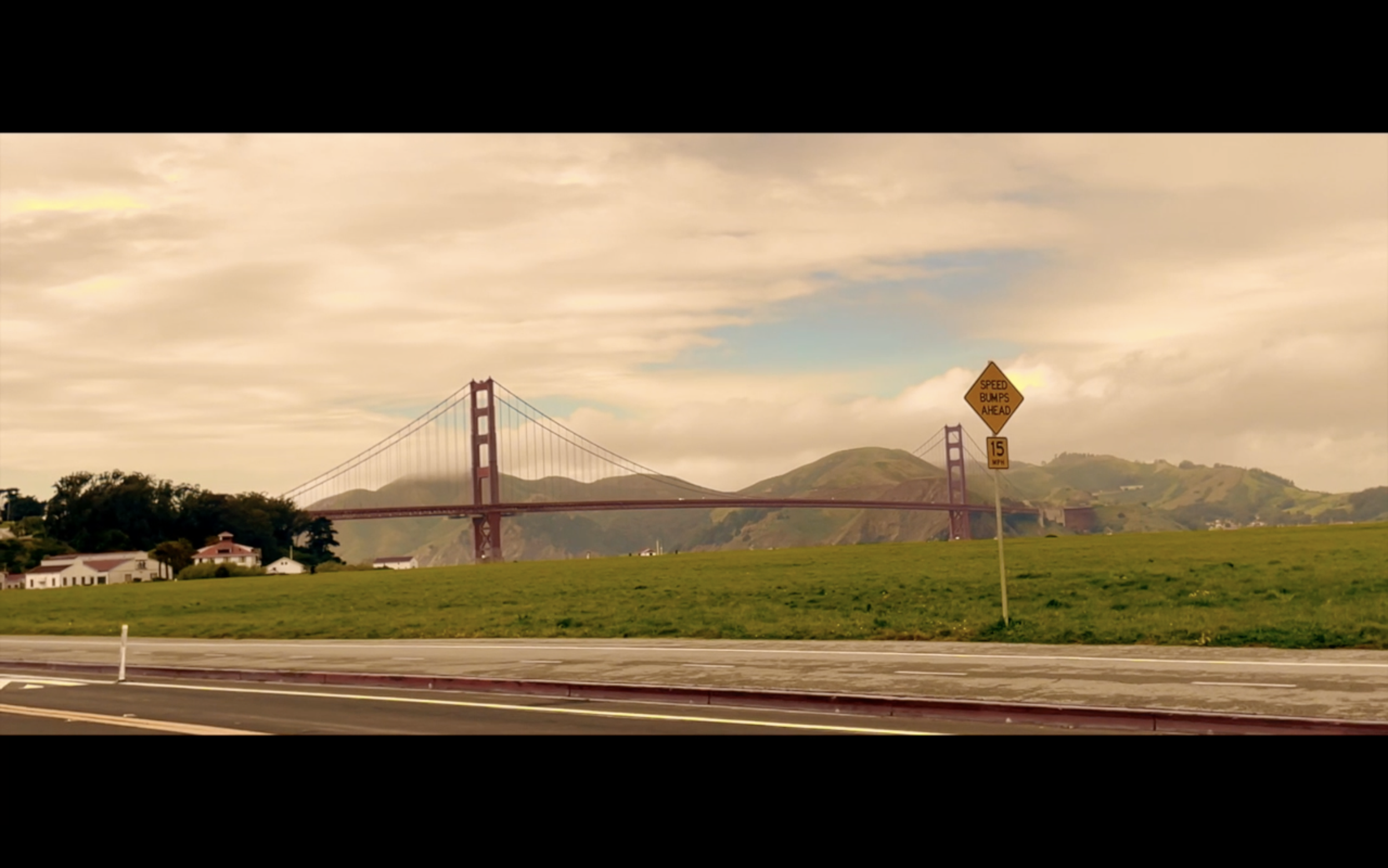 View of the Golden Gate Bridge in San Francisco with a speed bump warning sign in the foreground, grassy areas, hills, and partly cloudy sky