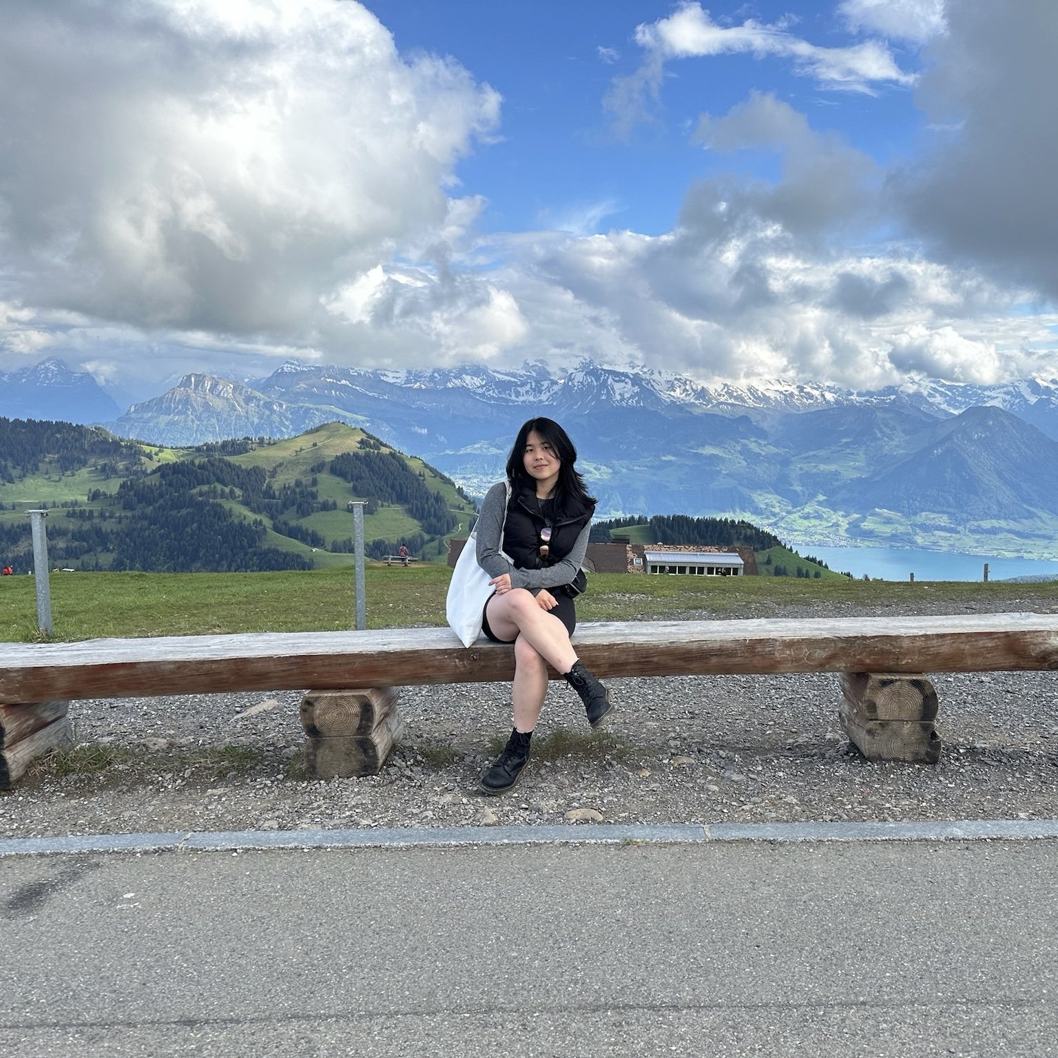 A woman sitting on a wooden bench outdoors with a mountain landscape in the background, including snow-capped peaks and green hills.