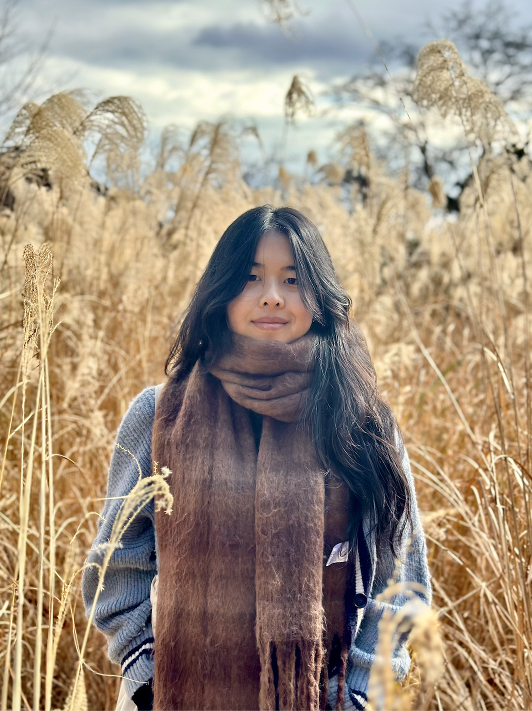 A woman with long dark hair and a brown scarf standing in a field of tall, golden dry grass or reeds on a cloudy day.