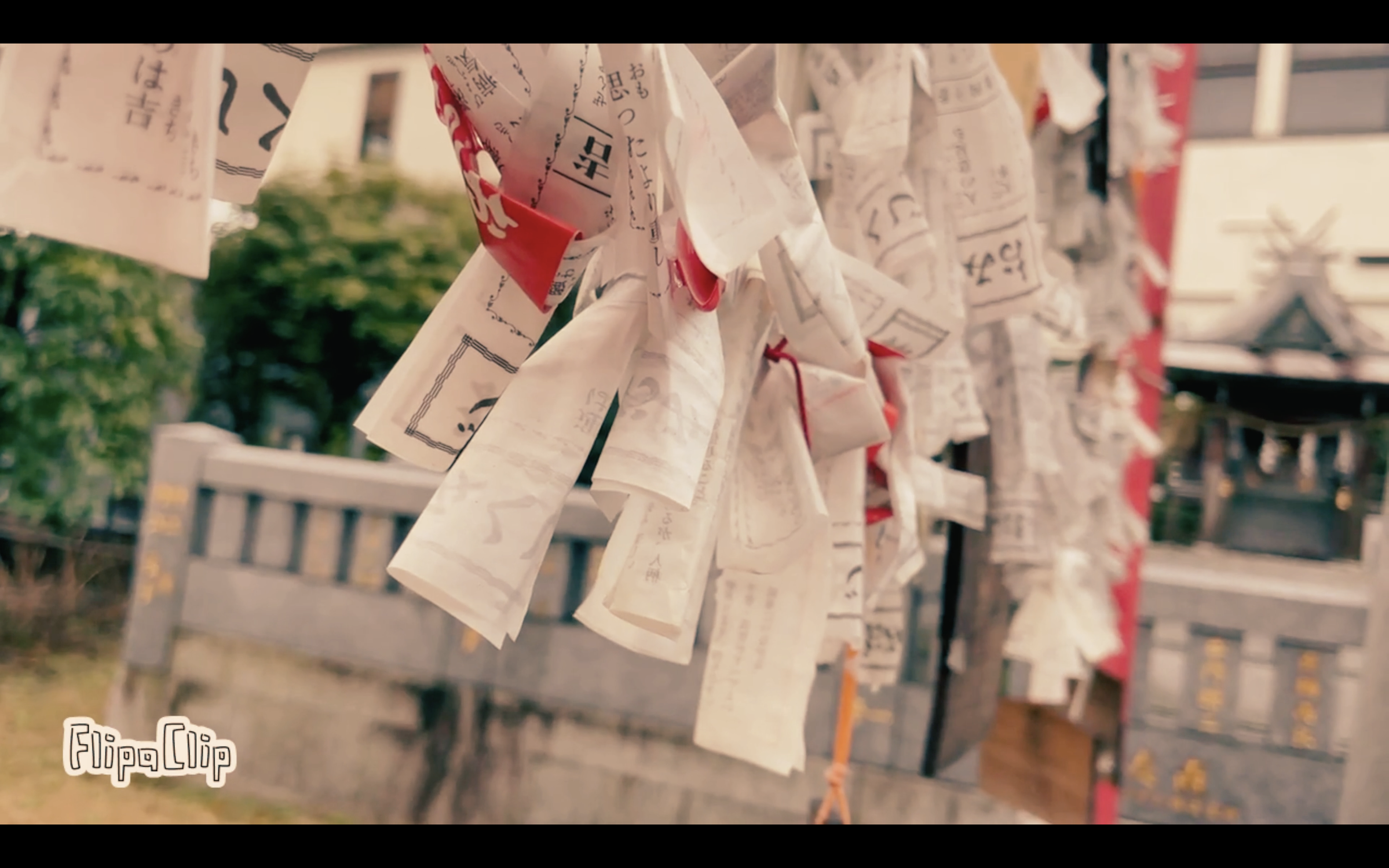 Close-up of strips of paper with Japanese characters tied to a wooden frame at a shrine, with a traditional building visible in the background.