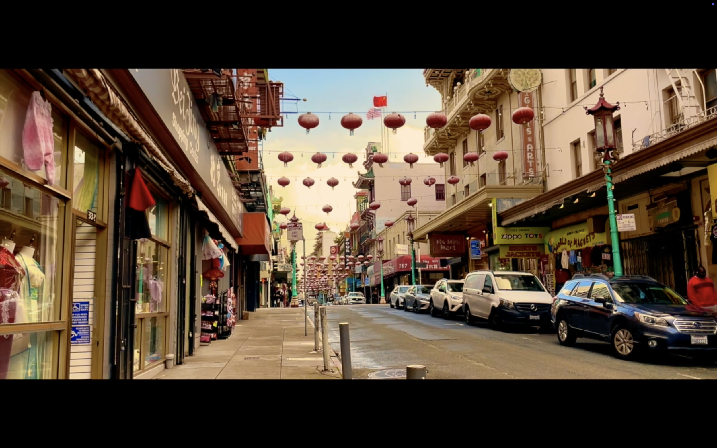 Street view of Chinatown with red lanterns hanging above, storefronts, and parked cars.