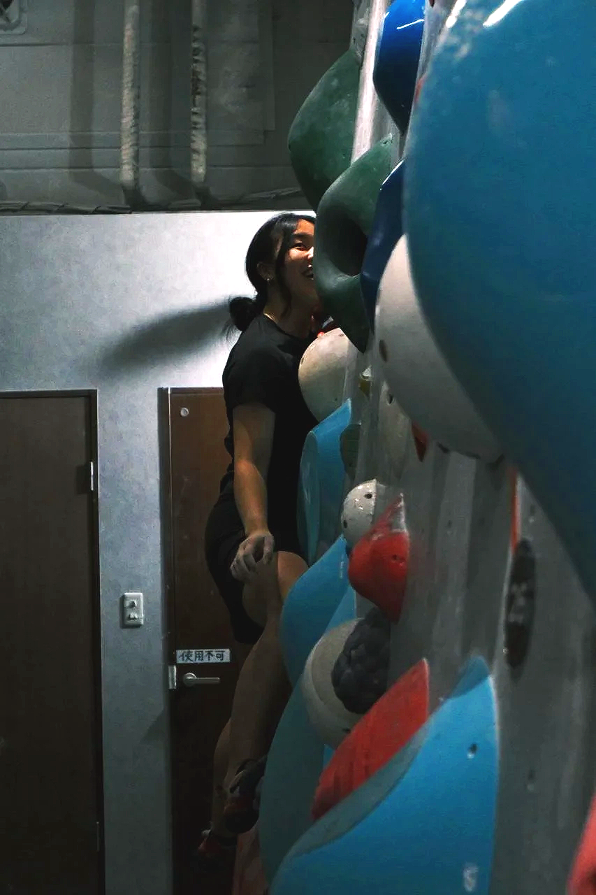A woman climbing an indoor rock climbing wall, smiling, in a black t-shirt and shorts, with various colorful climbing holds.