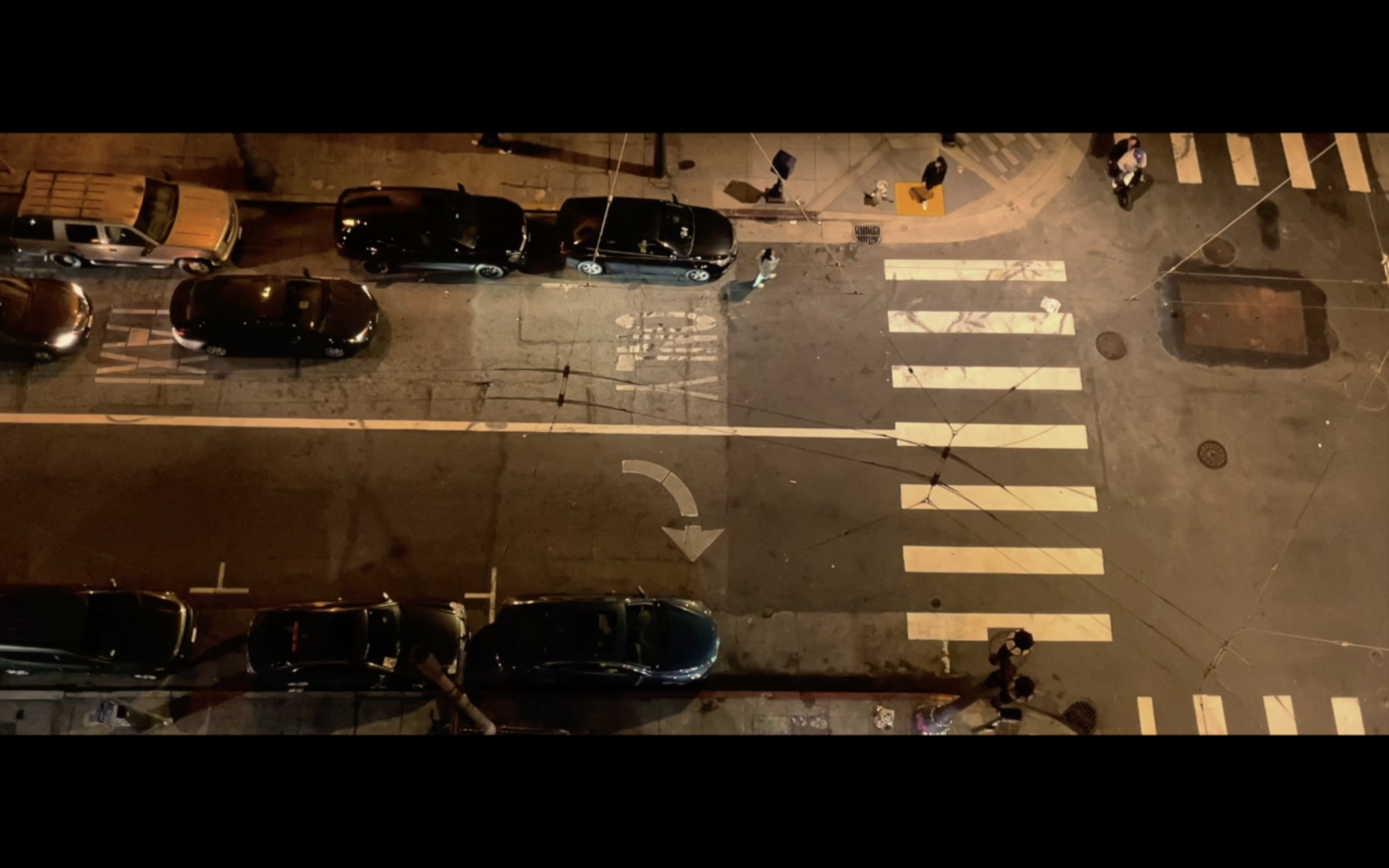 Nighttime aerial view of a city street showing parked cars, pedestrians crossing a crosswalk, and street markings, illuminated by streetlights.