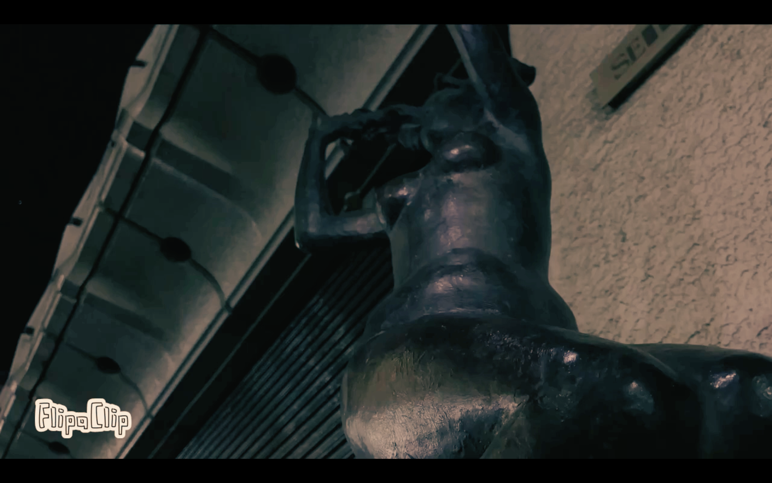 Close-up of a dark metallic humanoid sculpture with muscular details, leaning against or resting on a surface, with surrounding objects and a textured wall in the background.