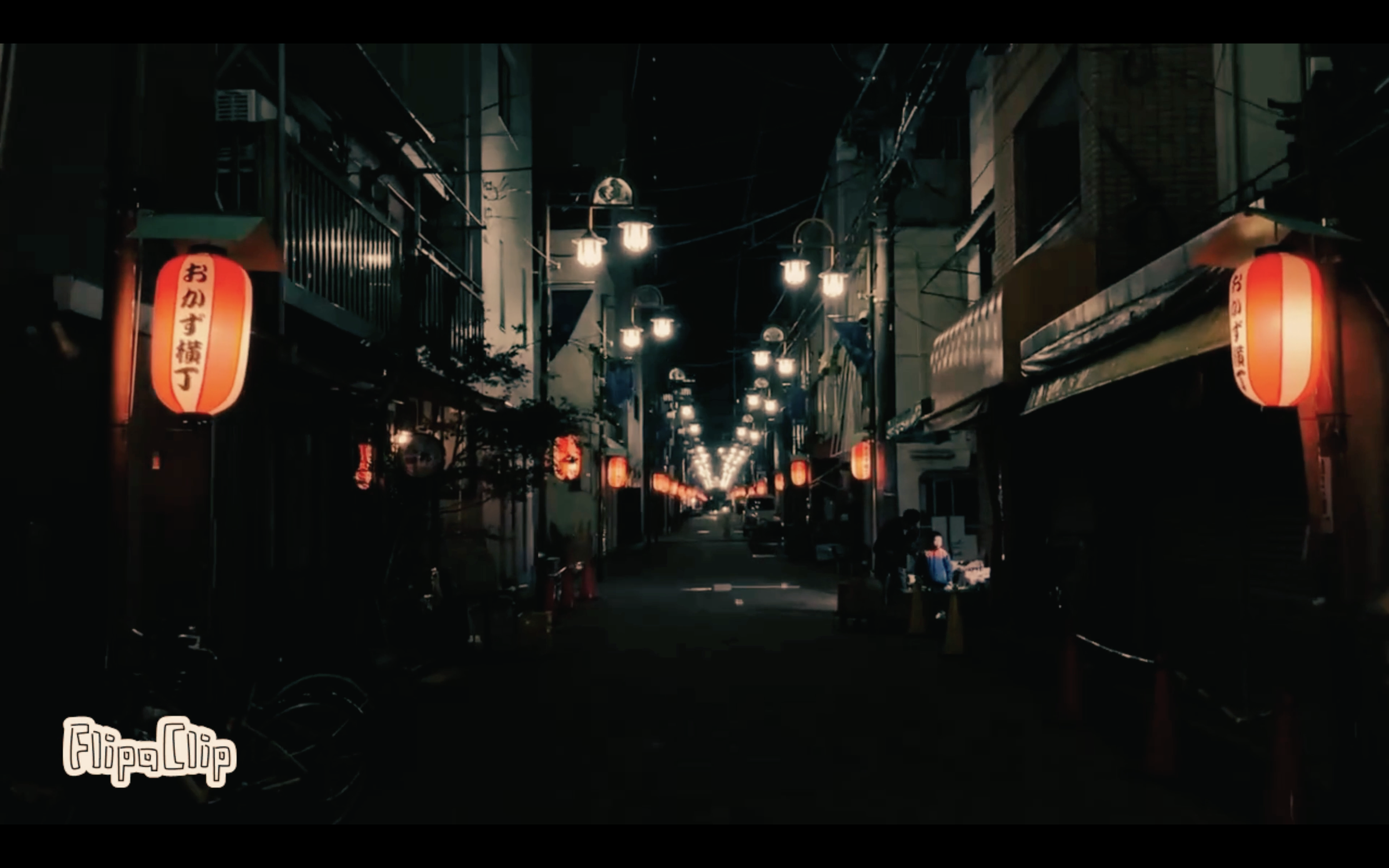 Nighttime street scene in a Japanese shopping district, illuminated by hanging paper lanterns and street lamps, with some people seen on the sidewalk.