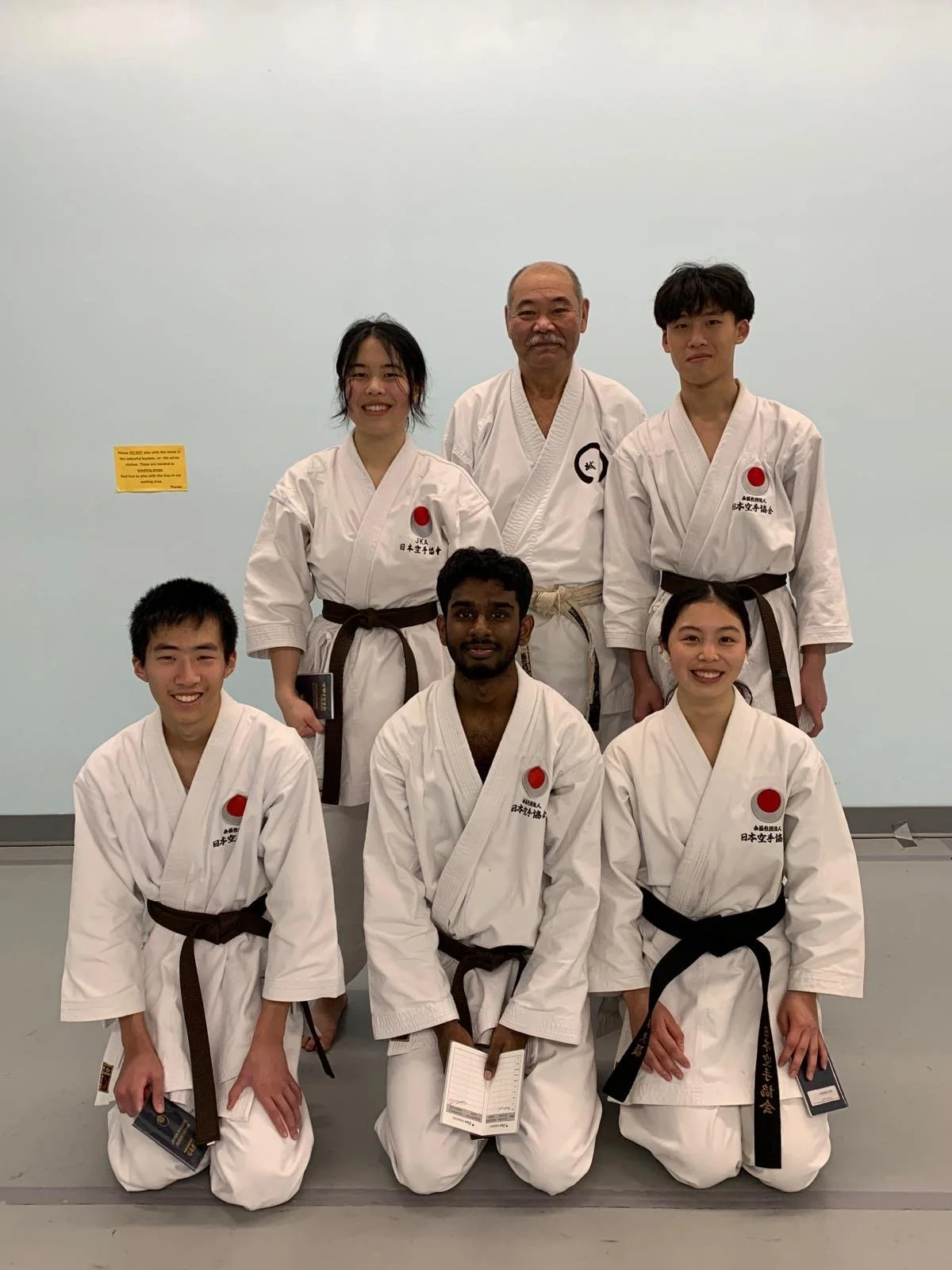 Group of seven martial artists wearing traditional karate uniforms with brown and black belts, posing for a photo in a dojo. They are kneeling and standing in two rows, with a plain wall background.