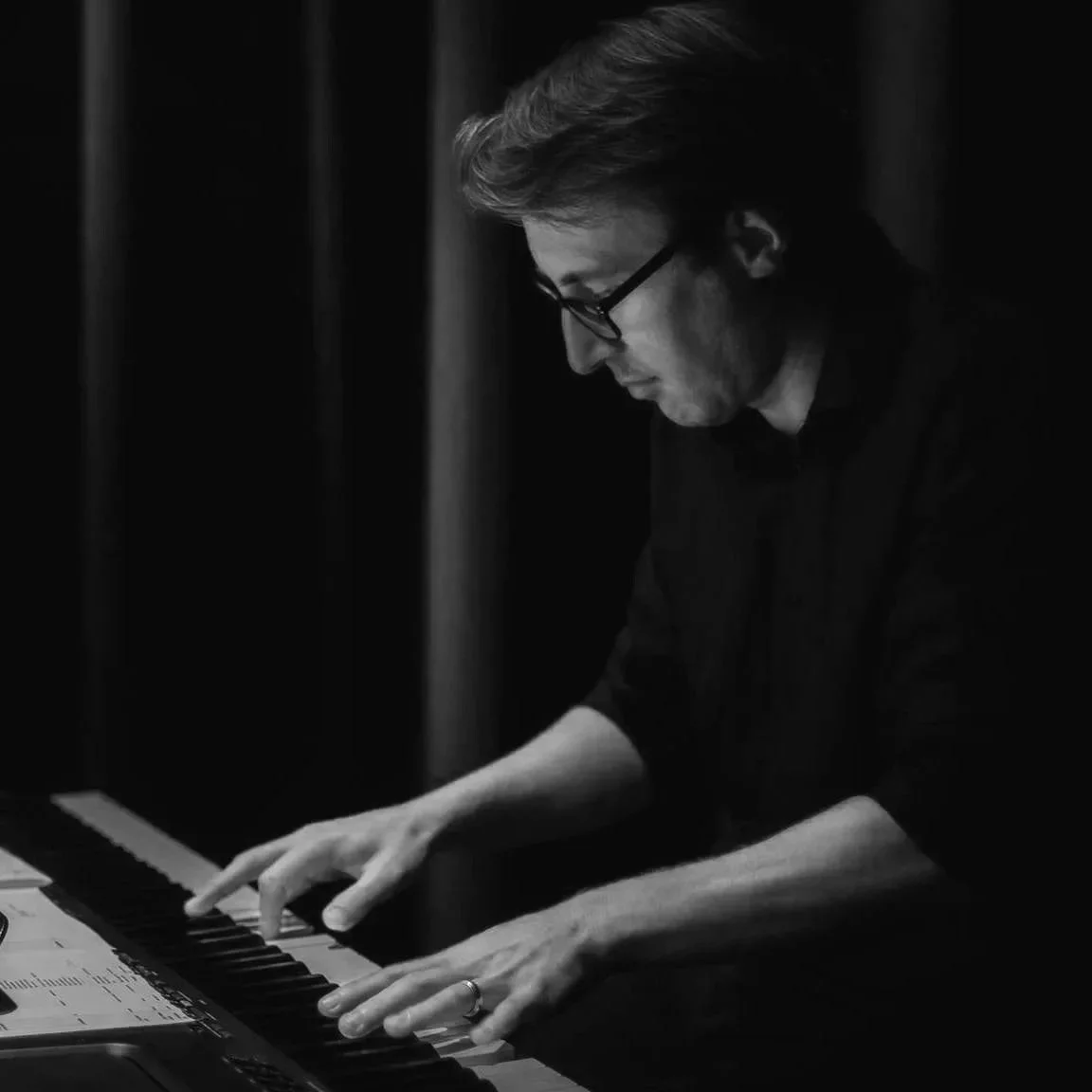 A person wearing glasses, playing the piano, focused on the keys, black and white photograph.