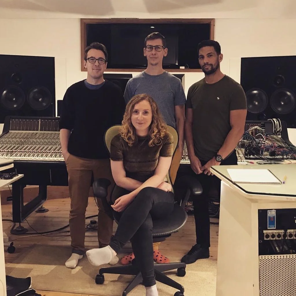 Four young adults in a recording studio, three men standing behind a woman seated in a rolling chair, surrounded by sound equipment and speakers.