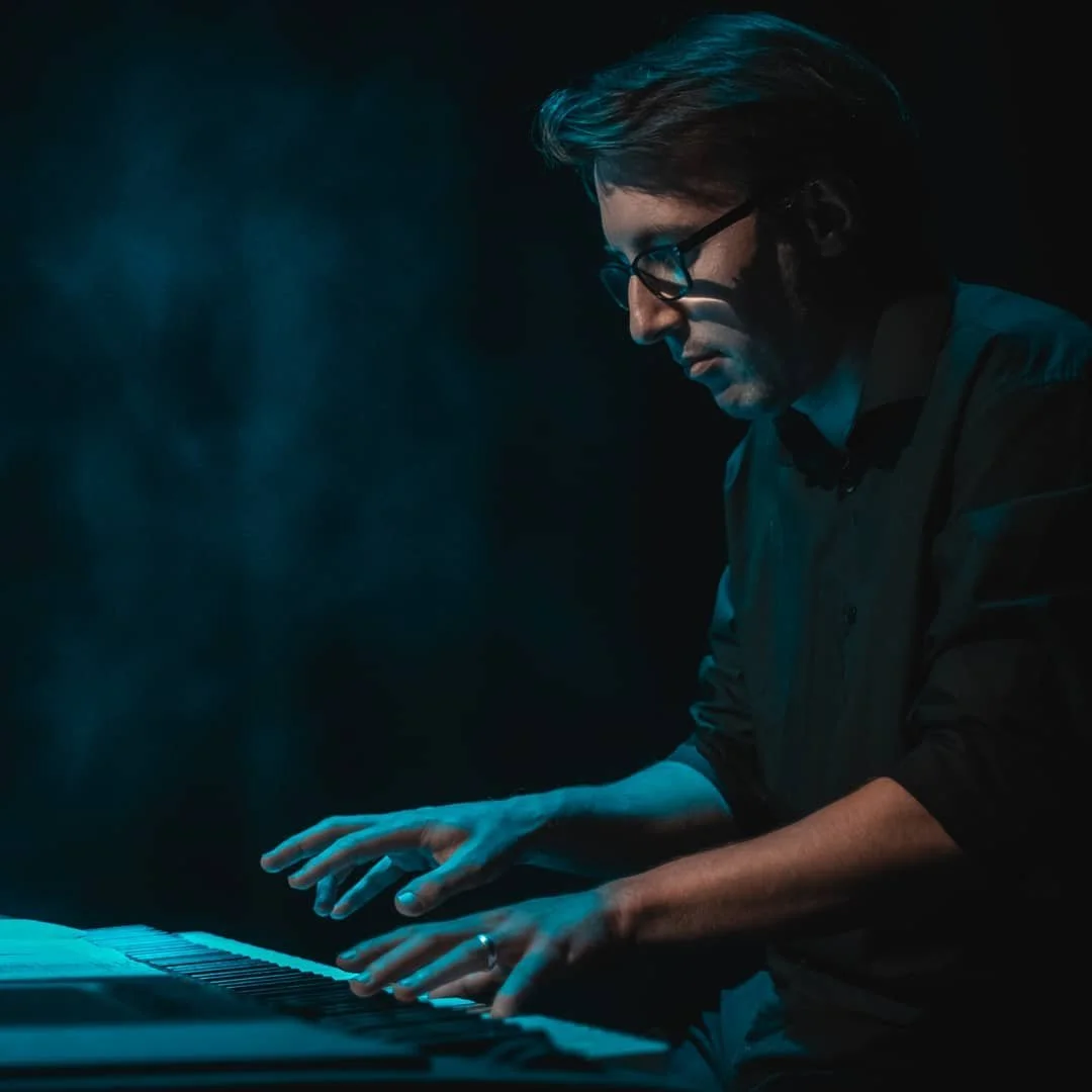 A person with glasses playing a keyboard piano in a dark setting with blue lighting.