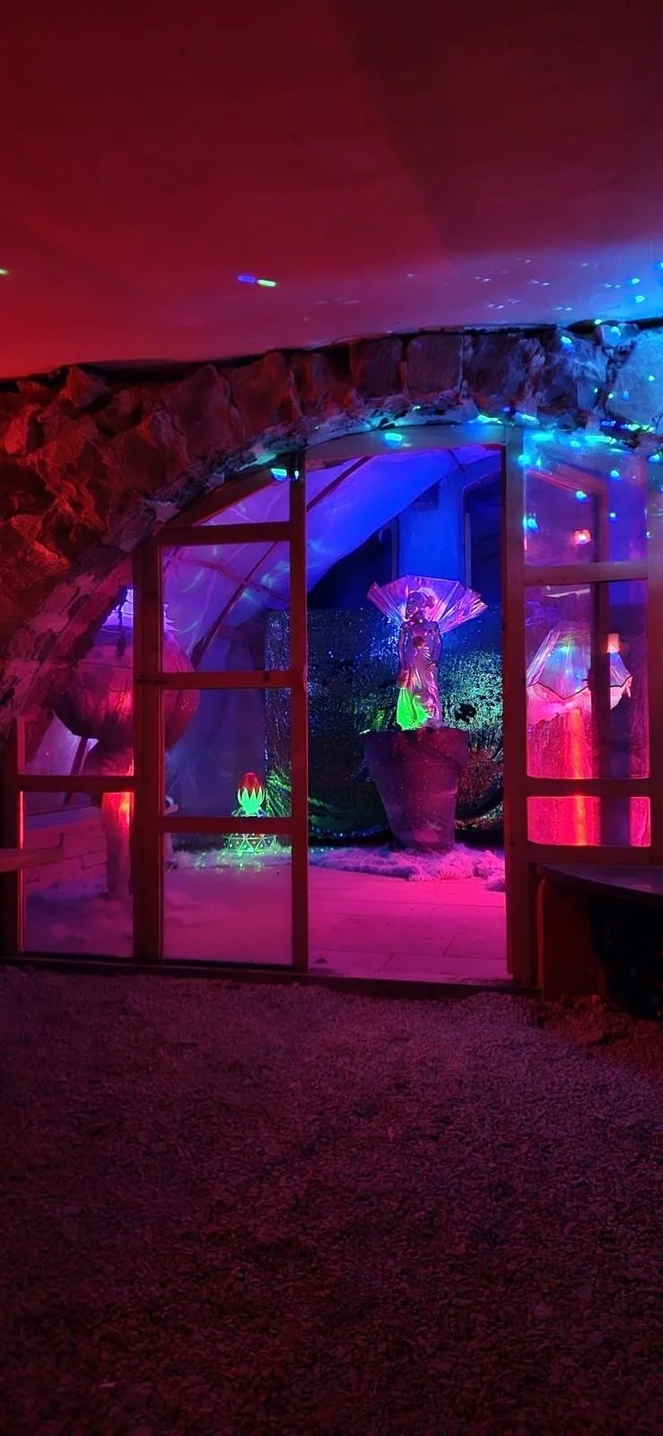 Bunte, leuchtende Dekoration in einer Höhle, mit einer Figur in einer Vase im Zentrum, umgeben von leuchtenden Lichtern und Neonfarben.