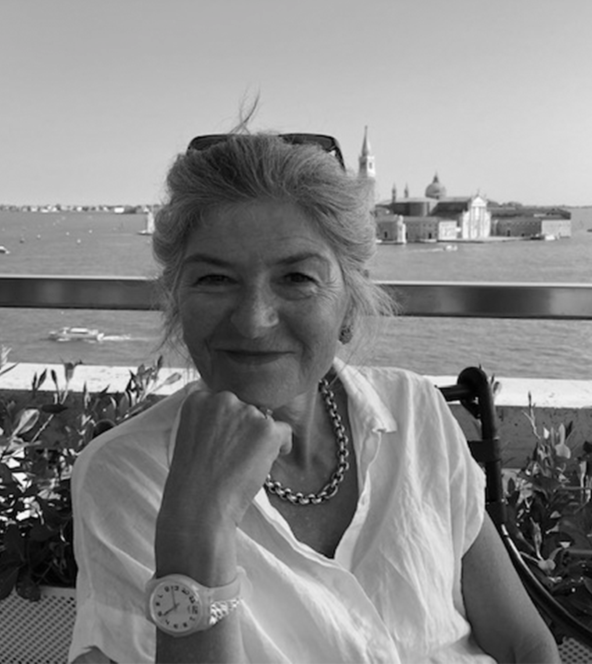 Eine ältere Frau mit grauem Haar, die auf einer Terrasse mit Blick auf eine Wasserlandschaft und eine Stadt im Hintergrund sitzt. Sie trägt eine weisse Bluse, eine Kette, eine Armbanduhr und Sonnenbrillen auf dem Kopf. Sie lächelt freundlich.