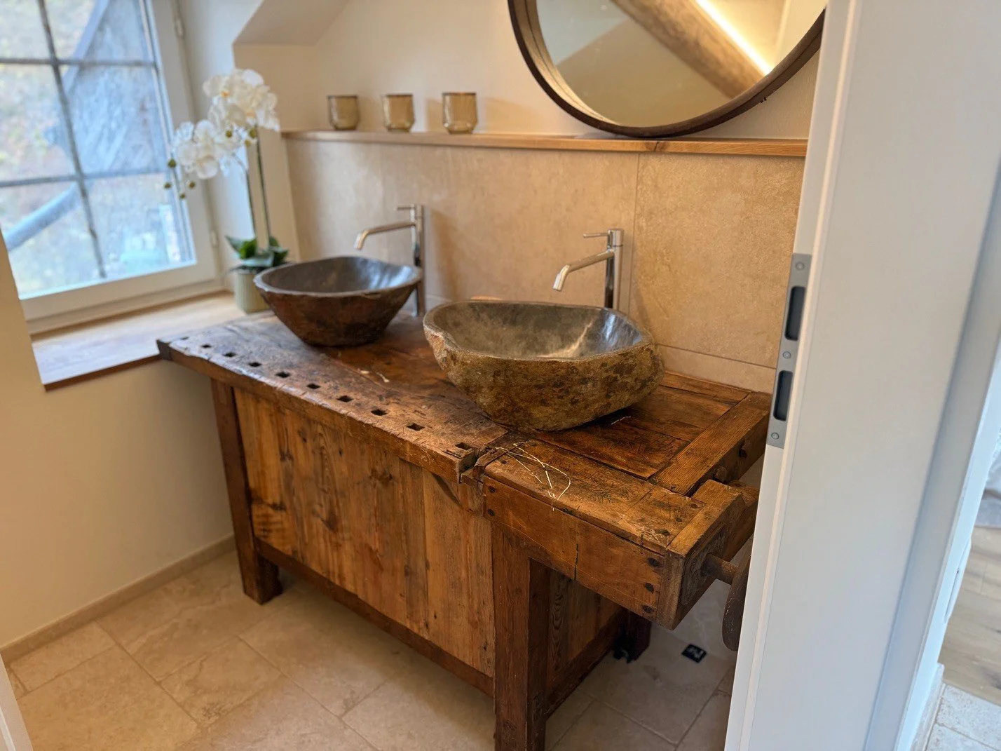 Das Badezimmer; zwei Steinschalen auf einem alten Holzwaschbecken, Wasserhähne darüber, neben einem Fenster mit weissen Blumen.