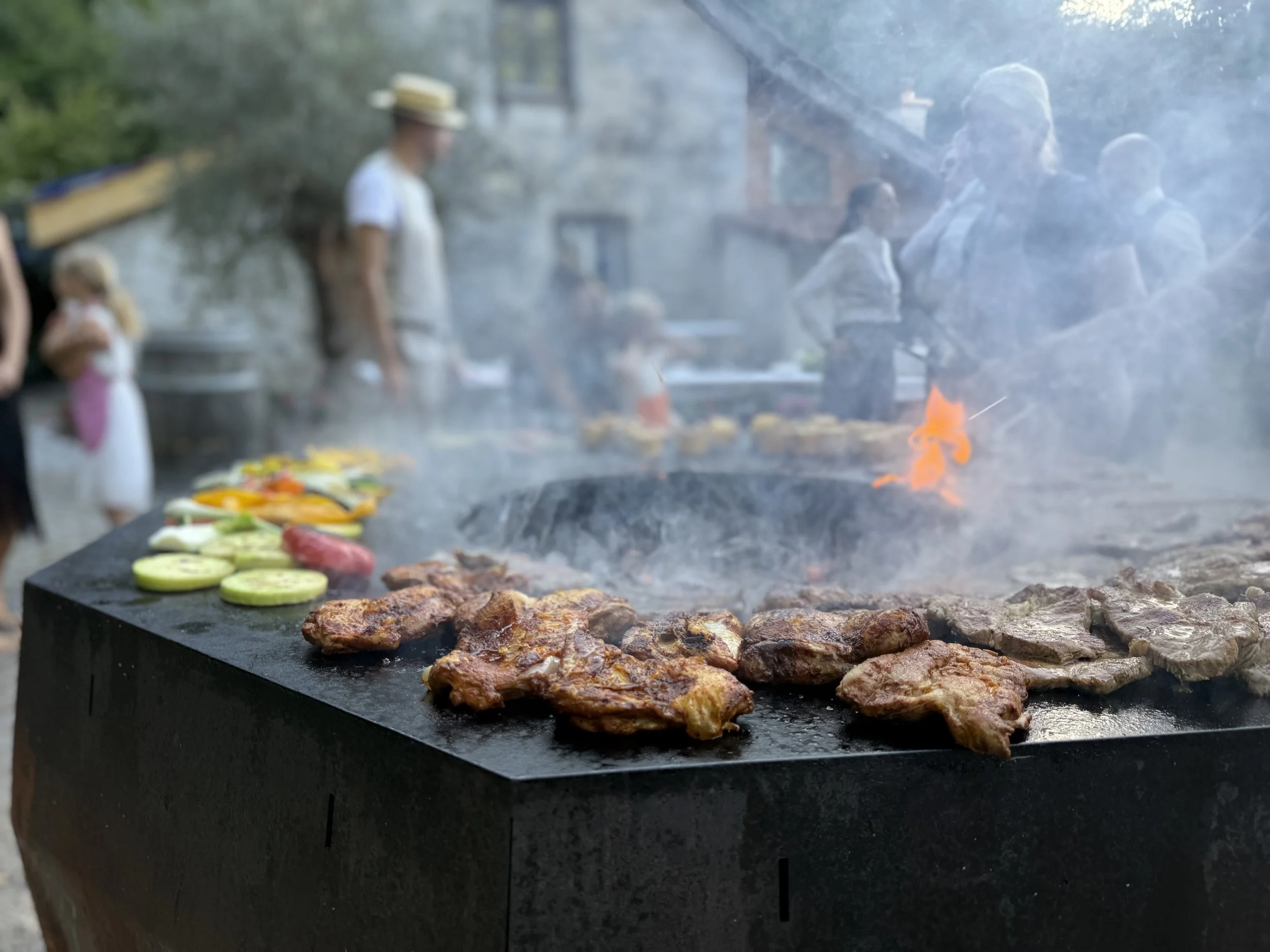 Grill mit Fleisch und Gemüse auf einem Feuerschalen-Grill, Menschen im Hintergrund bei einem Grillfest.