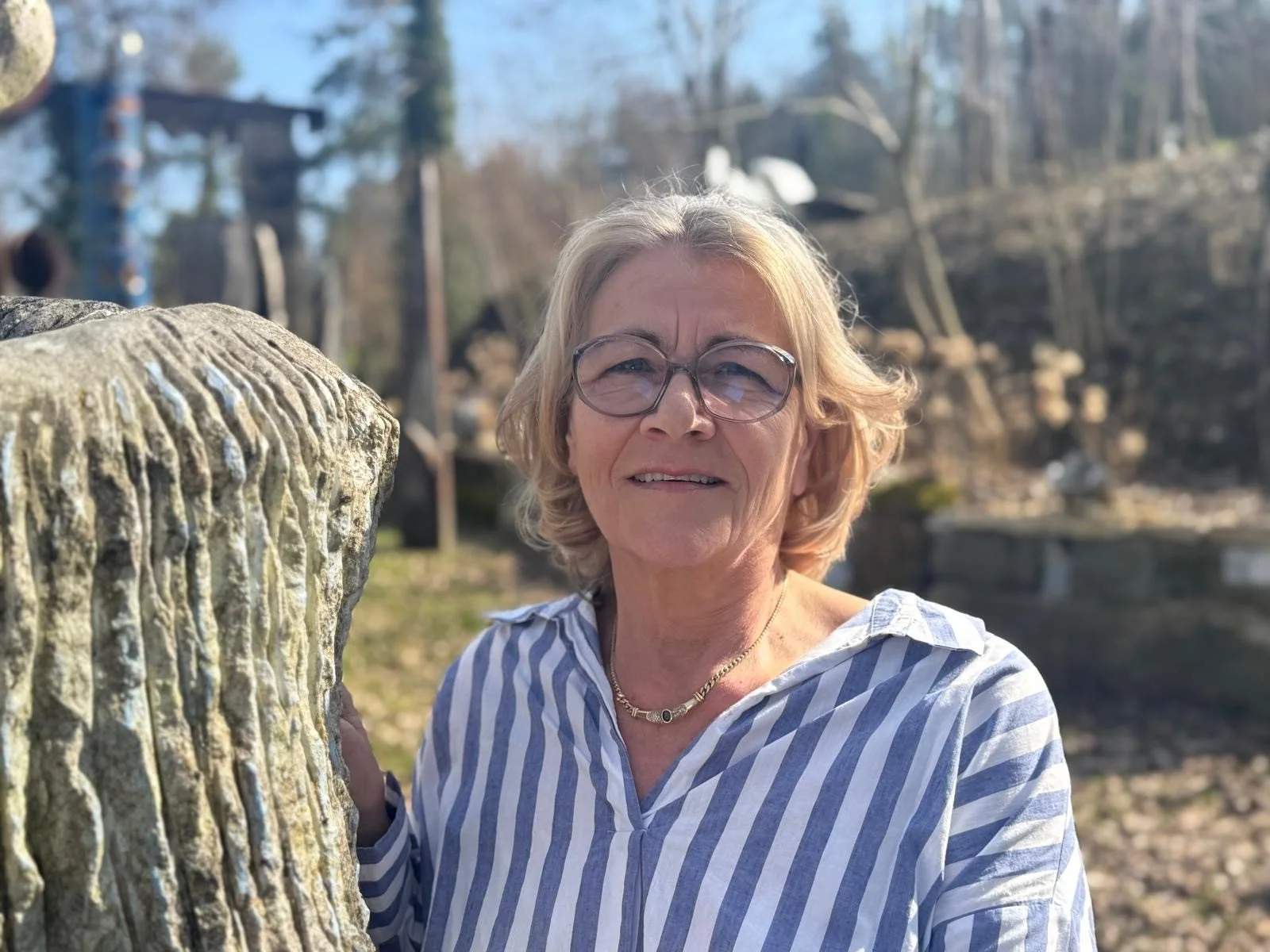 Renate Müller steht lächelnd im Freien, neben einem Baumstamm, im Hintergrund sind Bäume und ein Felsen sichtbar.