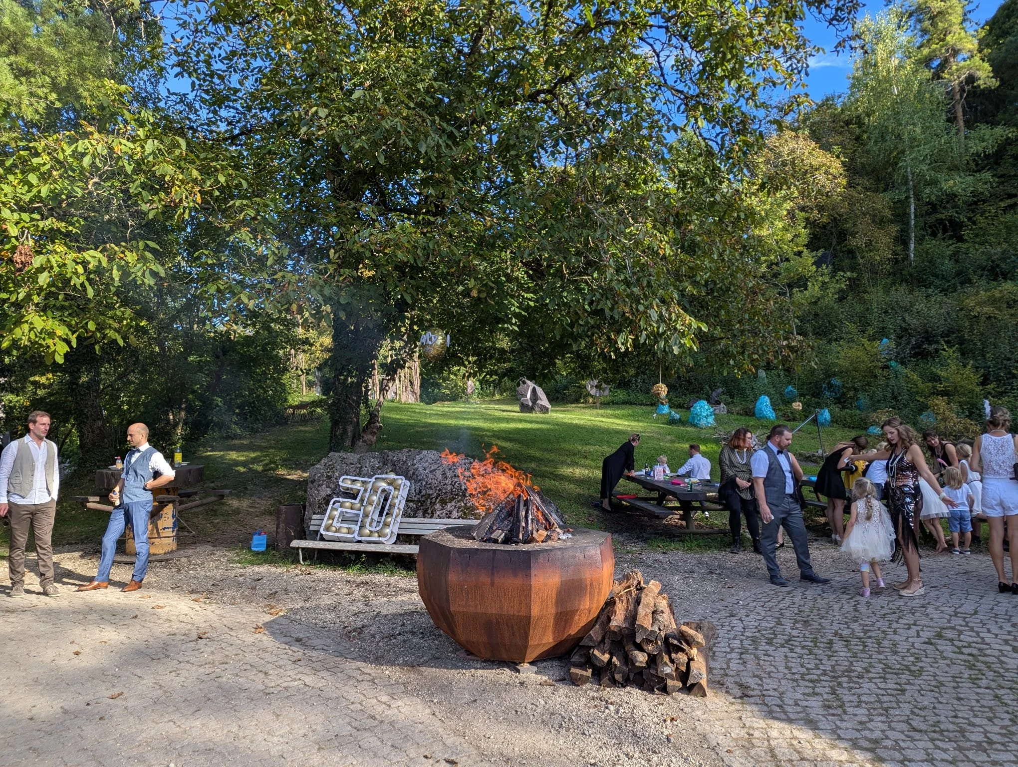 Ein Outdoor-Gartenfest mit Menschen, die um ein Lagerfeuer sitzen und feiern, dekoriert mit bunten Lampions und einer fröhlichen Atmosphäre.