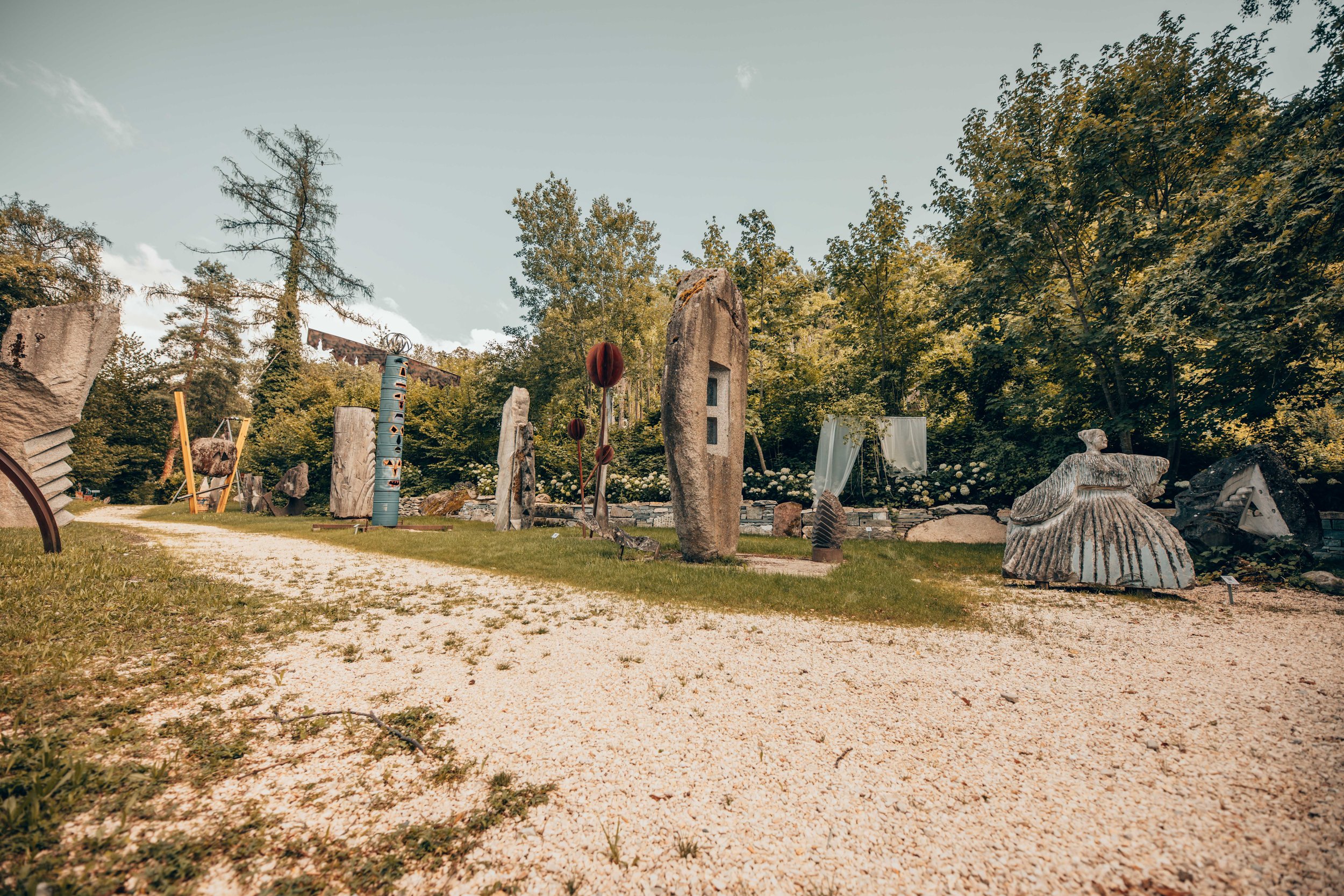 Skulpturengarten mit verschiedenen Kunstwerken aus Stein und Metall umgeben von Bäumen und Rasen, bei daylight.