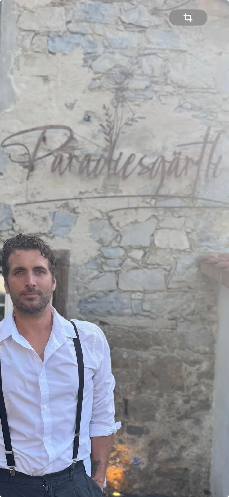 Thomas Müller, Inhaber des Paradiesgärtli steht vor einer alten Steinhinterwand mit Wandkunst, die einen Rosenstrauch und das Wort 'Paradiesgärtli' zeigt.