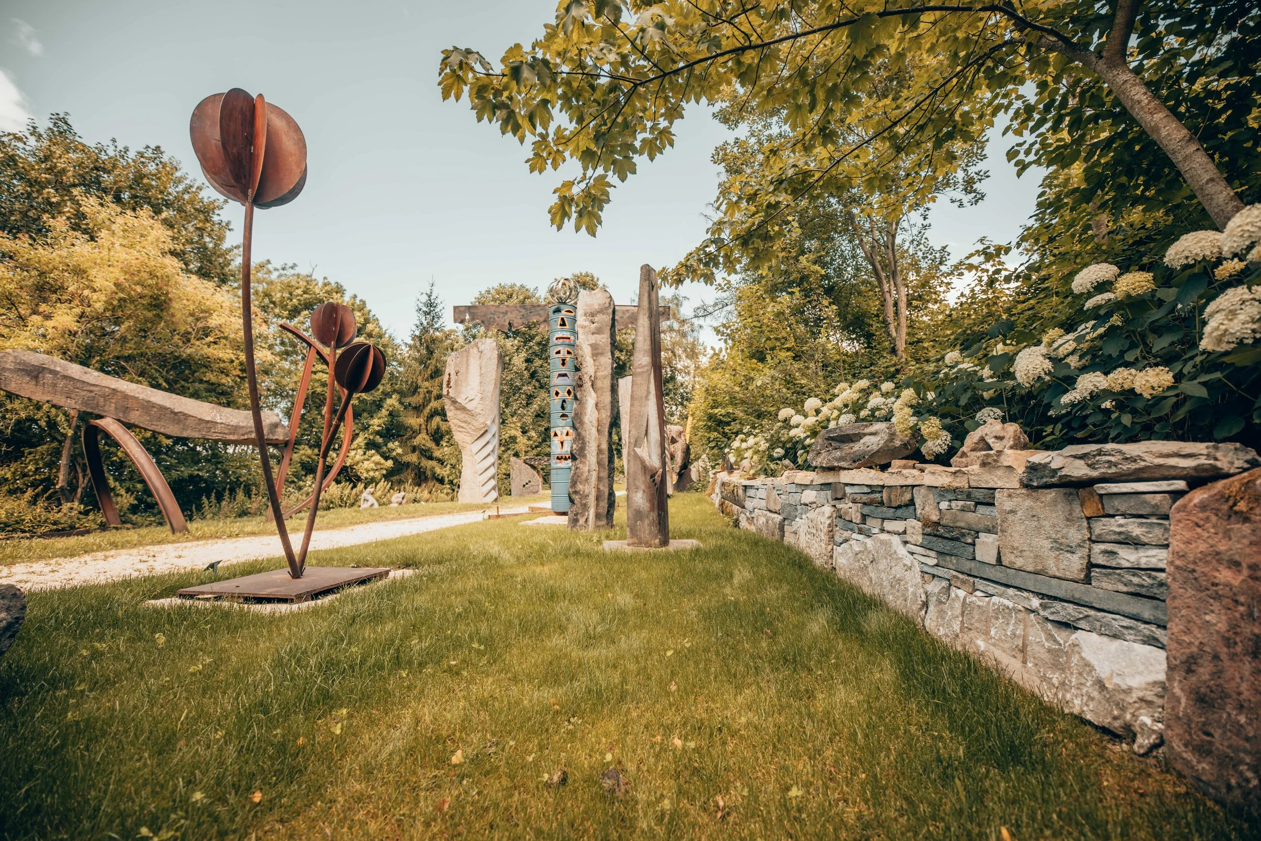 Skulpturengarten mit modernen Kunstwerken, Steinskulpturen, Metallskulpturen, Gartenweg, umliegende Bäume und weisse Blumen, Bäume im Hintergrund, sonniger Himmel.